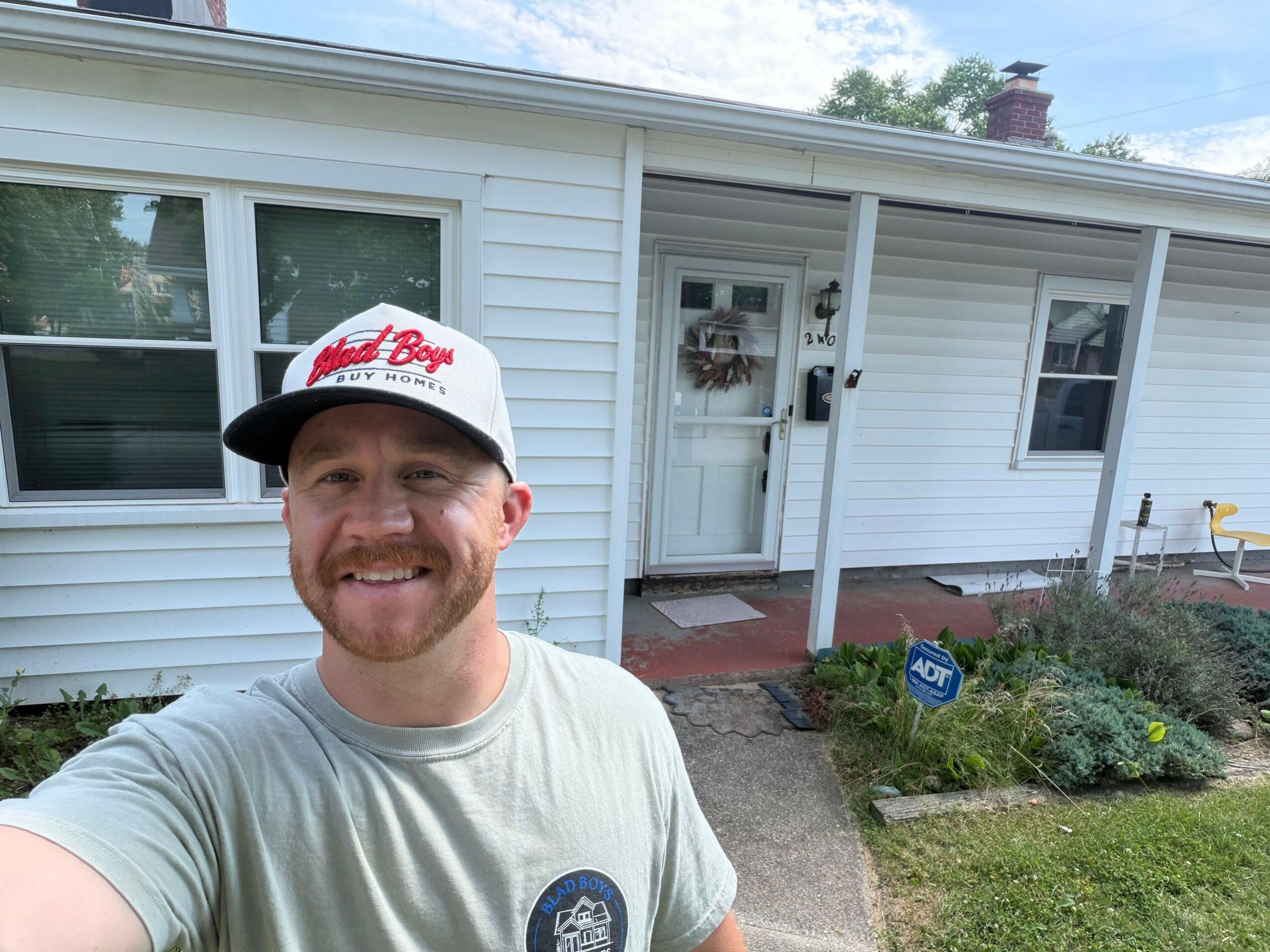 Man wearing a white cap and T-shirt smiling outside near a house with white siding and a garden.