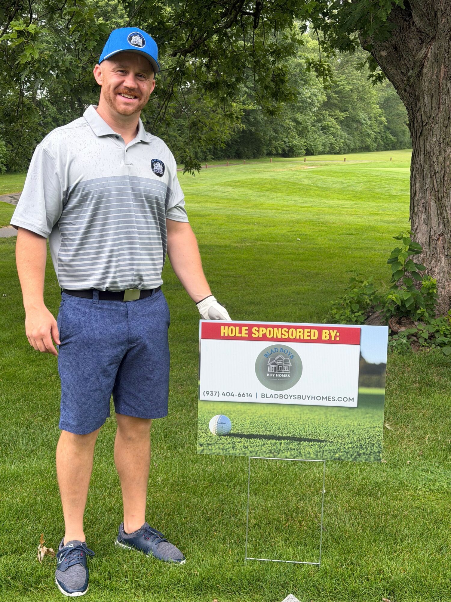 Young man in a striped shirt and shorts standing outdoors next to a sign on a golf course.