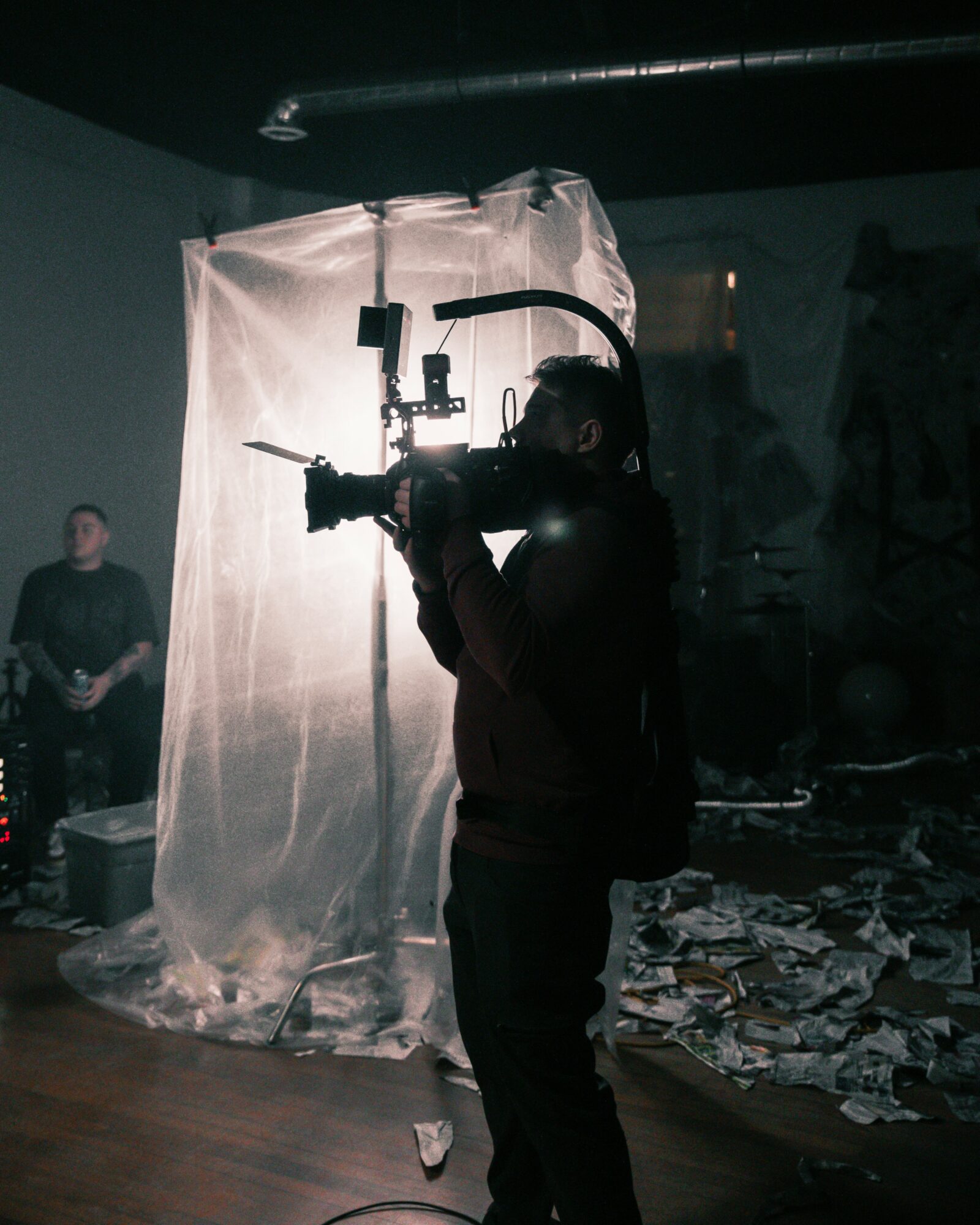 Person operating a large camera or filming equipment indoors, with a backdrop and scattered papers on the floor.