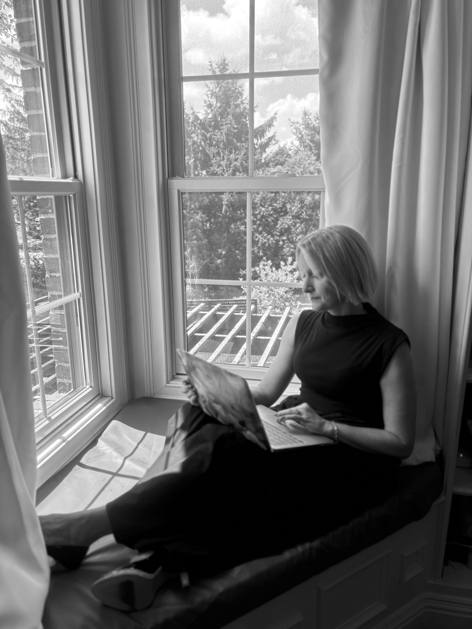 Woman sitting on window seat reading a book, with curtains and a view outside, in black and white.