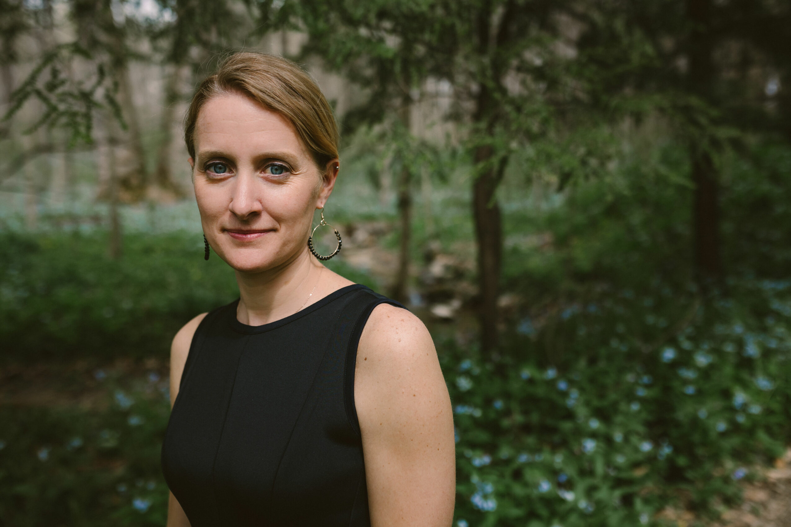 Woman with short hair in a sleeveless black top outdoors with trees and greenery in the background.