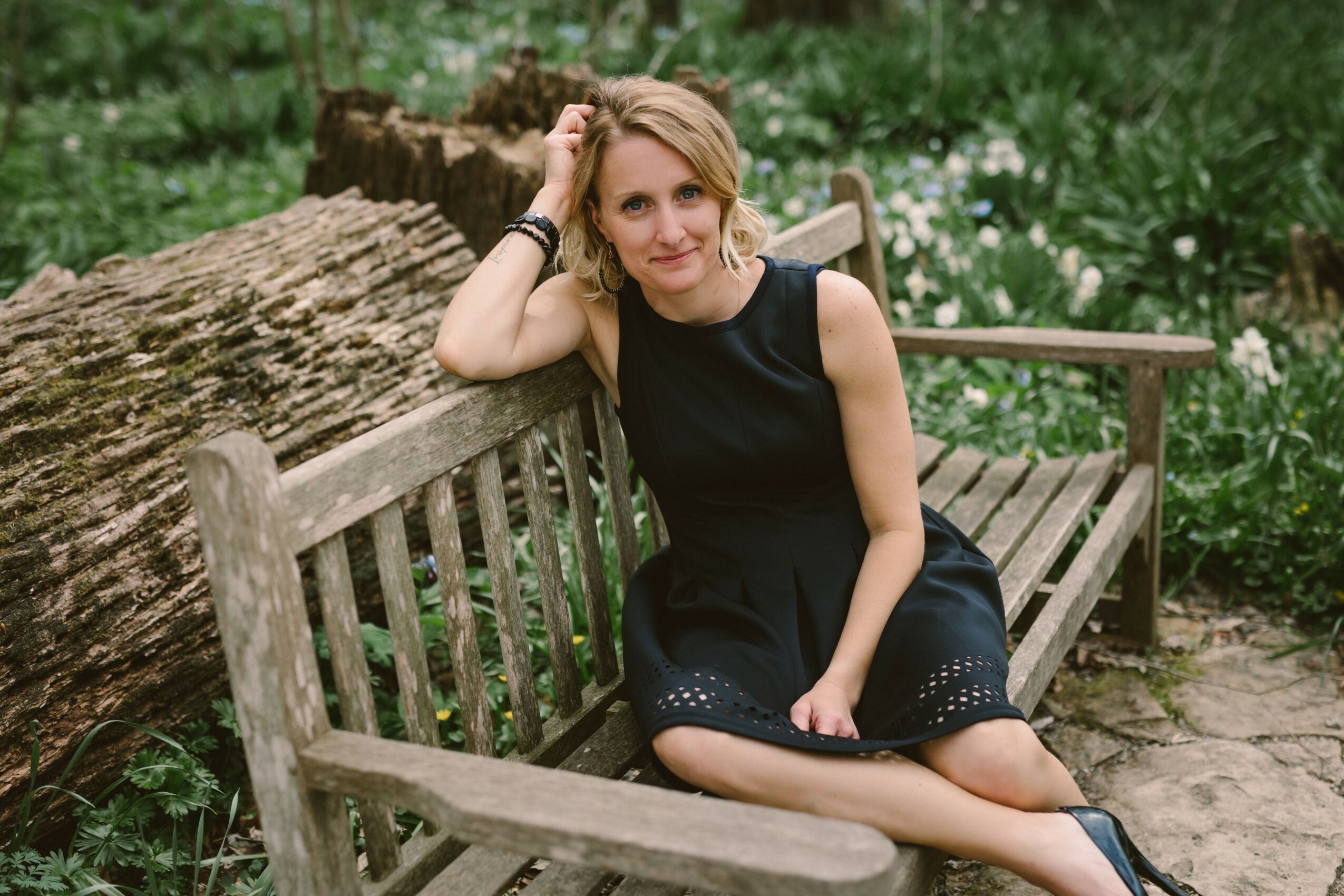 Woman sitting on a wooden bench outdoors, resting her head on her hand, surrounded by greenery and flowers.