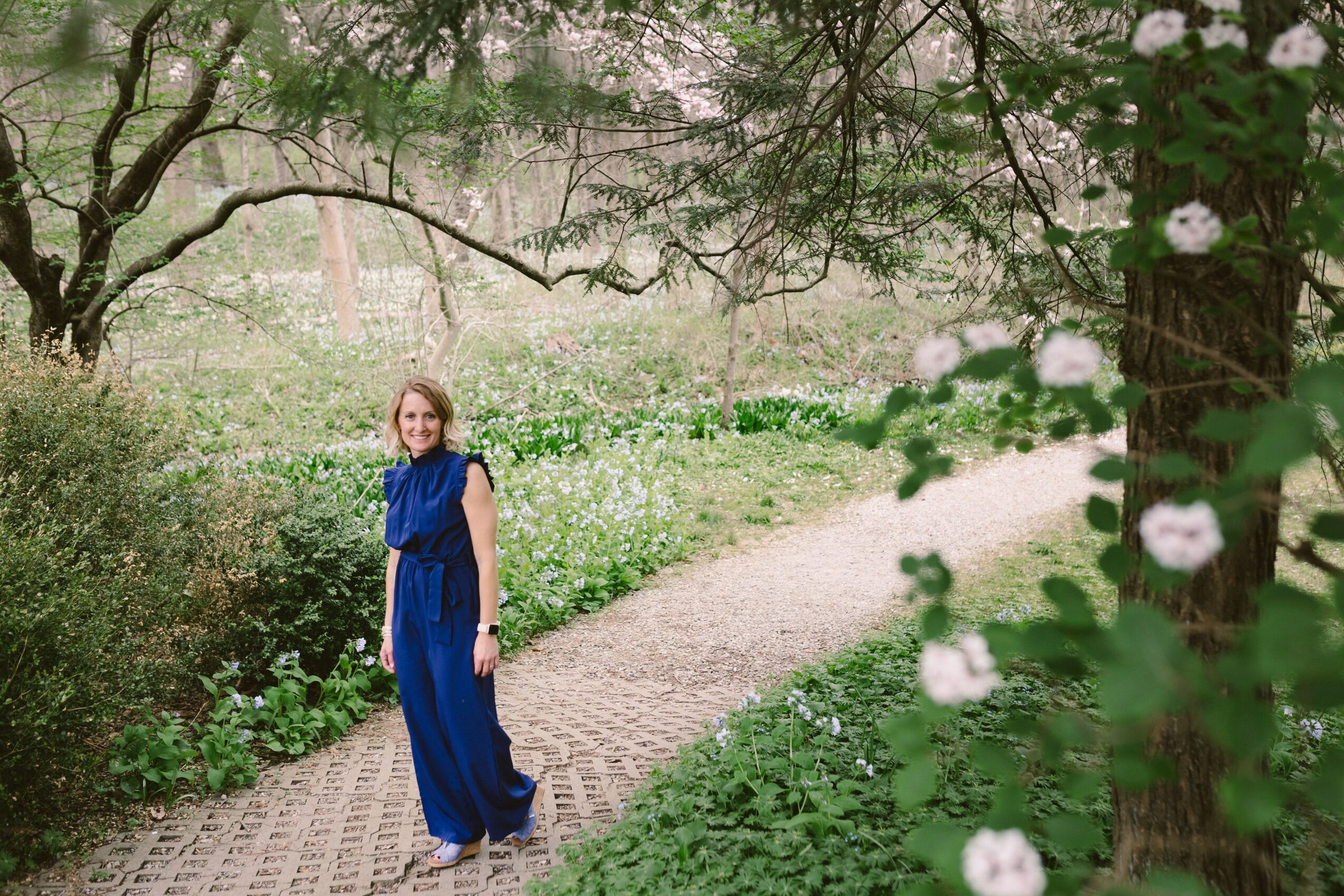 Young woman in blue dress walking on a garden path surrounded by greenery and flowering trees.