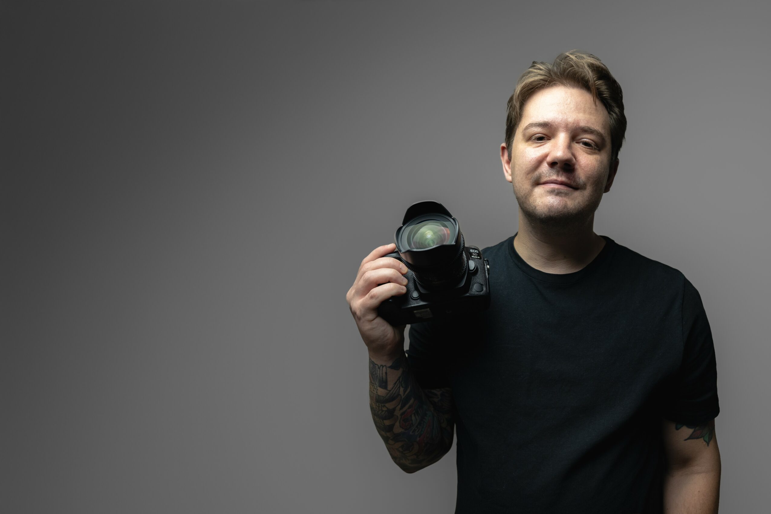 Young man with short hair holding a camera, smiling, wearing a black t-shirt, against a plain gray background.