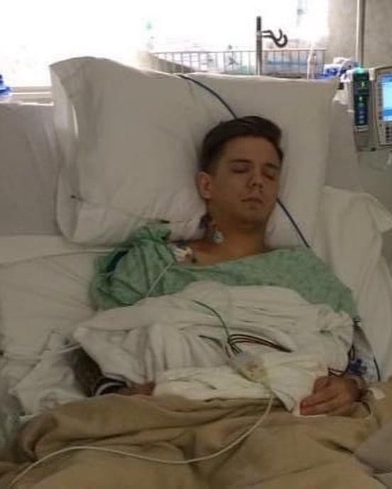 Young person with medical tubes and wires in a hospital bed, resting with eyes closed, pillow supporting head, medical equipment nearby.