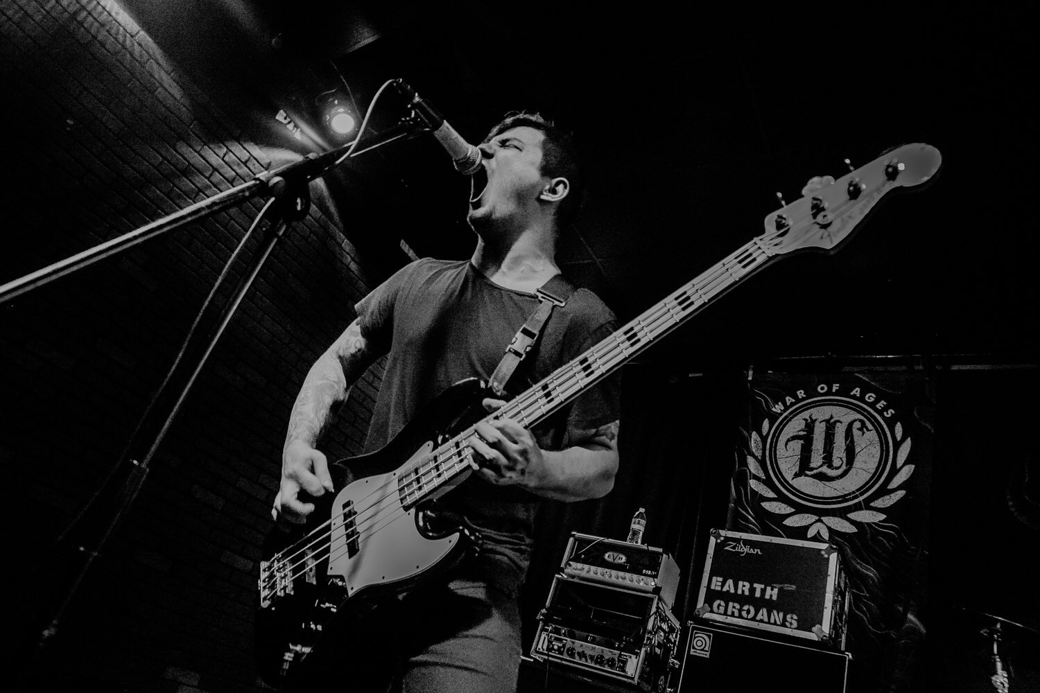 Musician playing bass guitar and singing into microphone on stage, with equipment and a sign in the background.