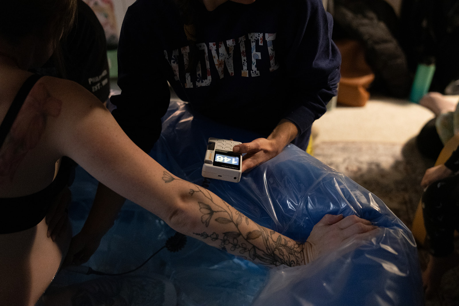 Person with tattooed arm reaching into a blue plastic bag, others around holding a phone, outdoor setting