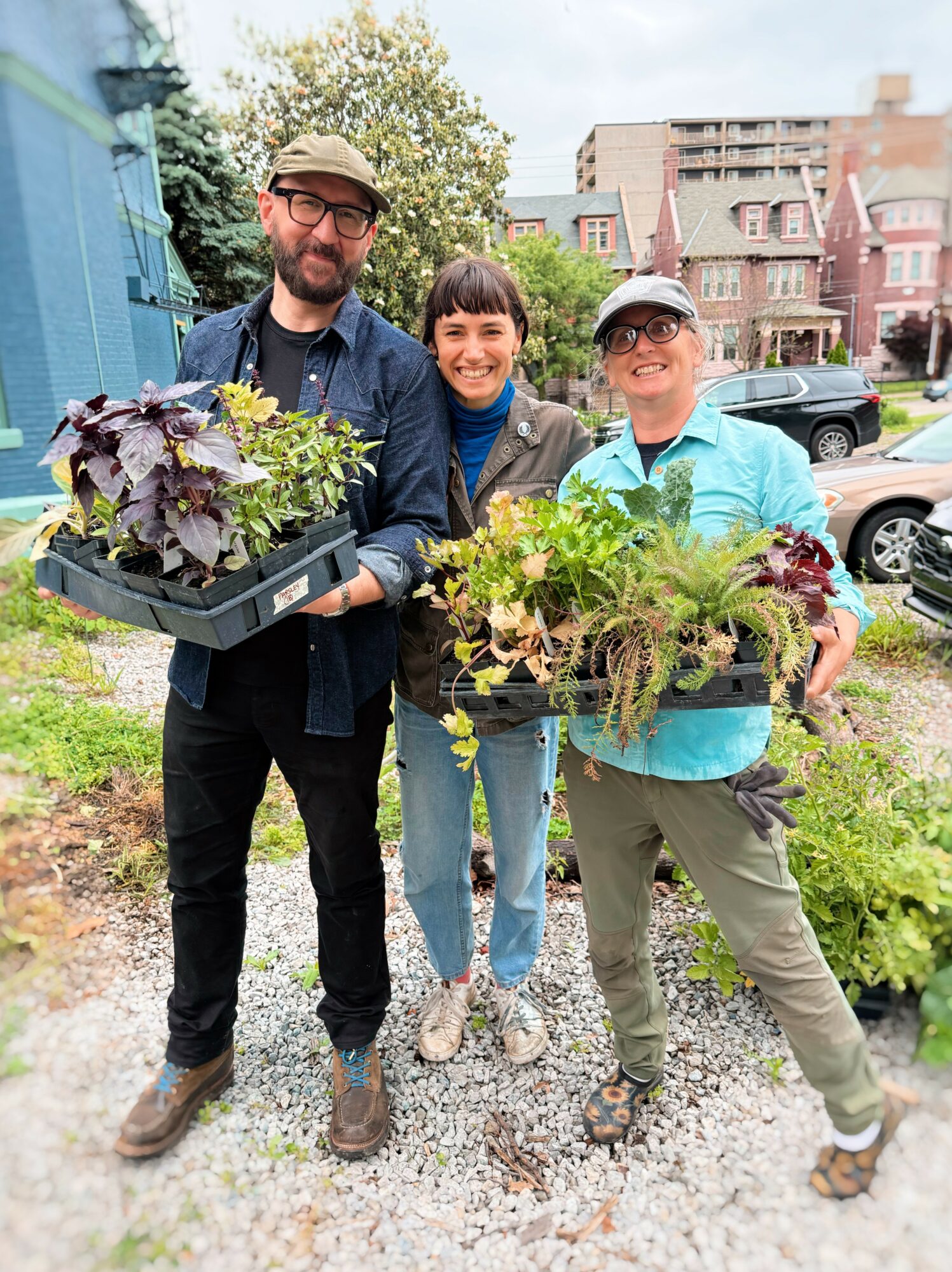 Three people holding trays of plants outdoors, smiling, with buildings and trees in the background.