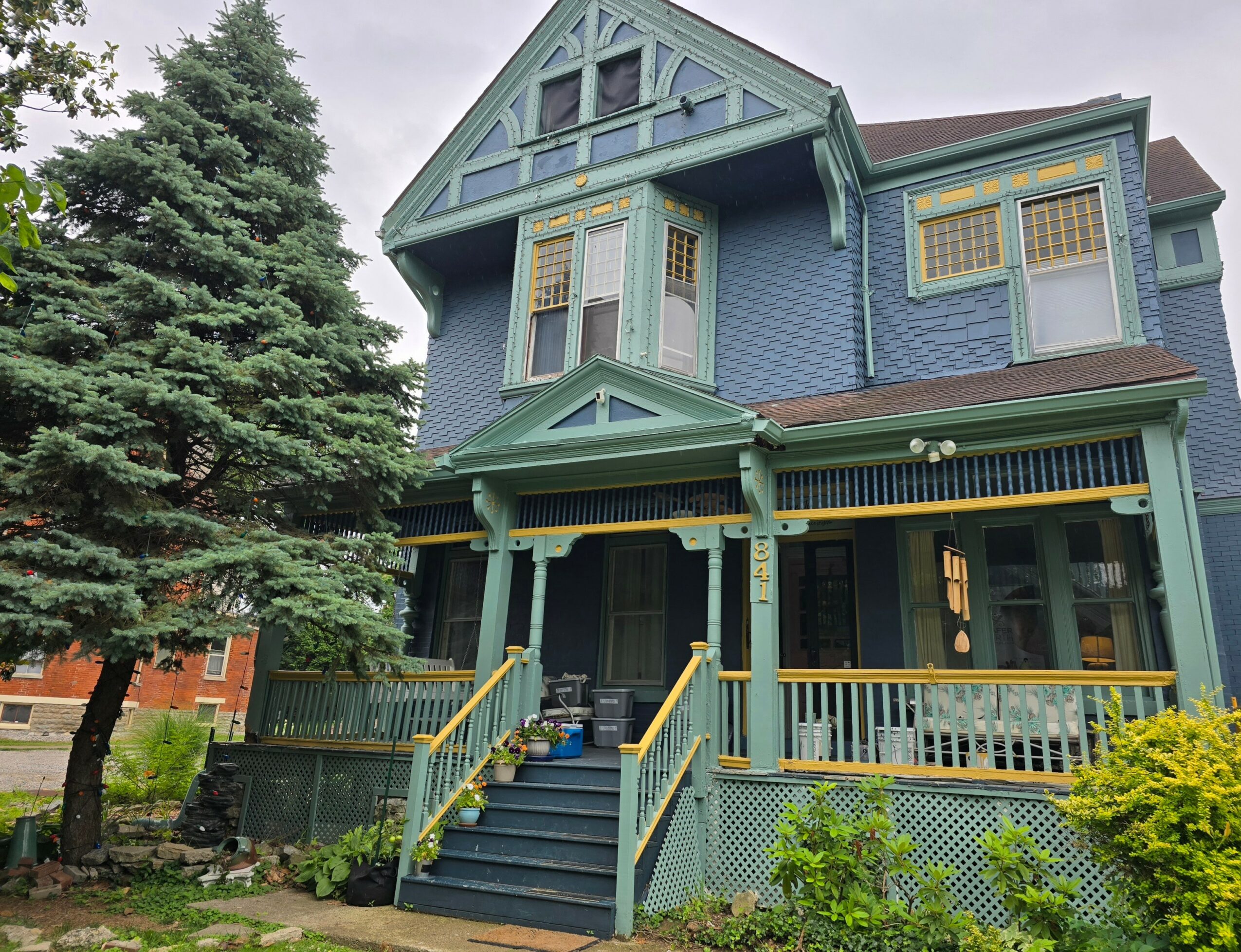 Three-story blue house with a front porch, surrounded by trees and greenery.