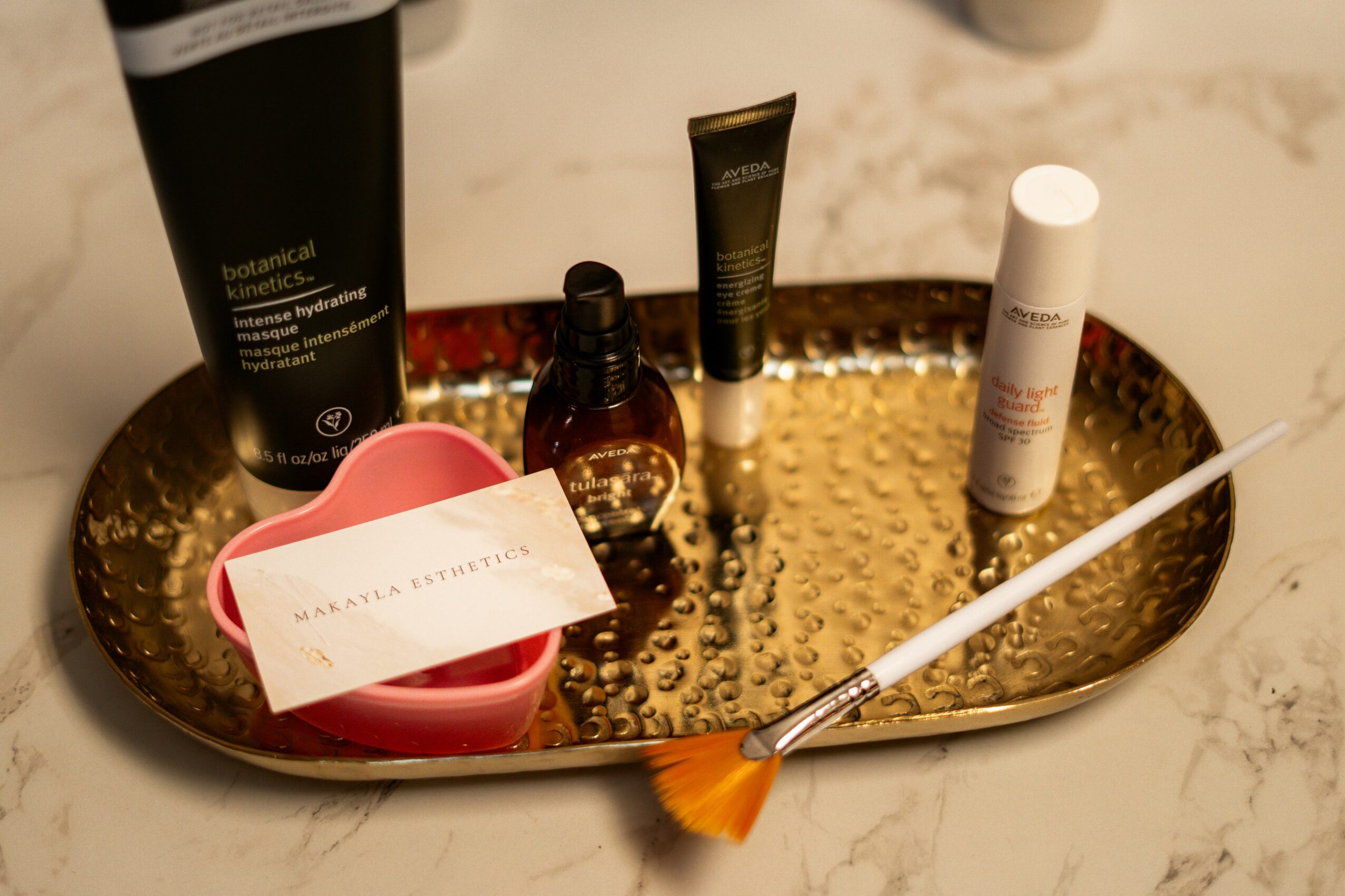 Gold tray with skincare products, a pink container, a card, and a brush on a marble surface.