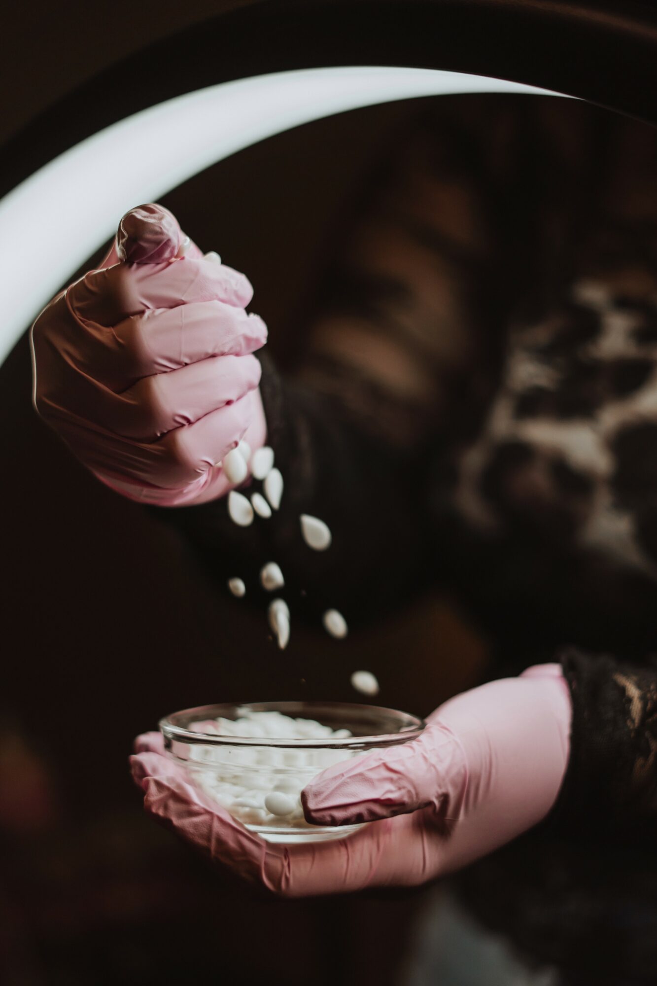 Person wearing pink gloves pouring white pills into a glass container, with a dark background and blurred pattern.