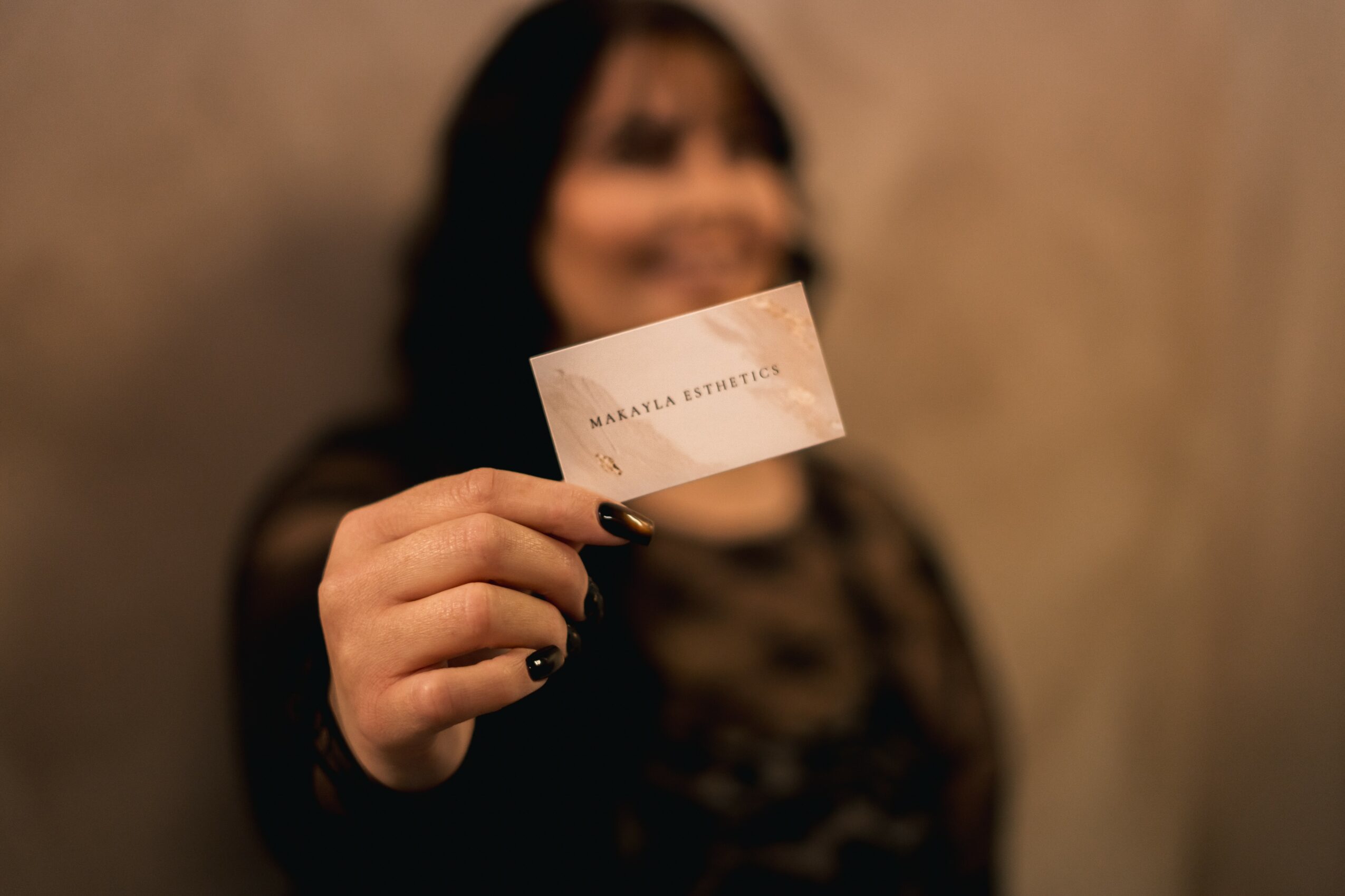 Person holding a white card with text, blurred background, dark clothing, and dark hair, smiling.