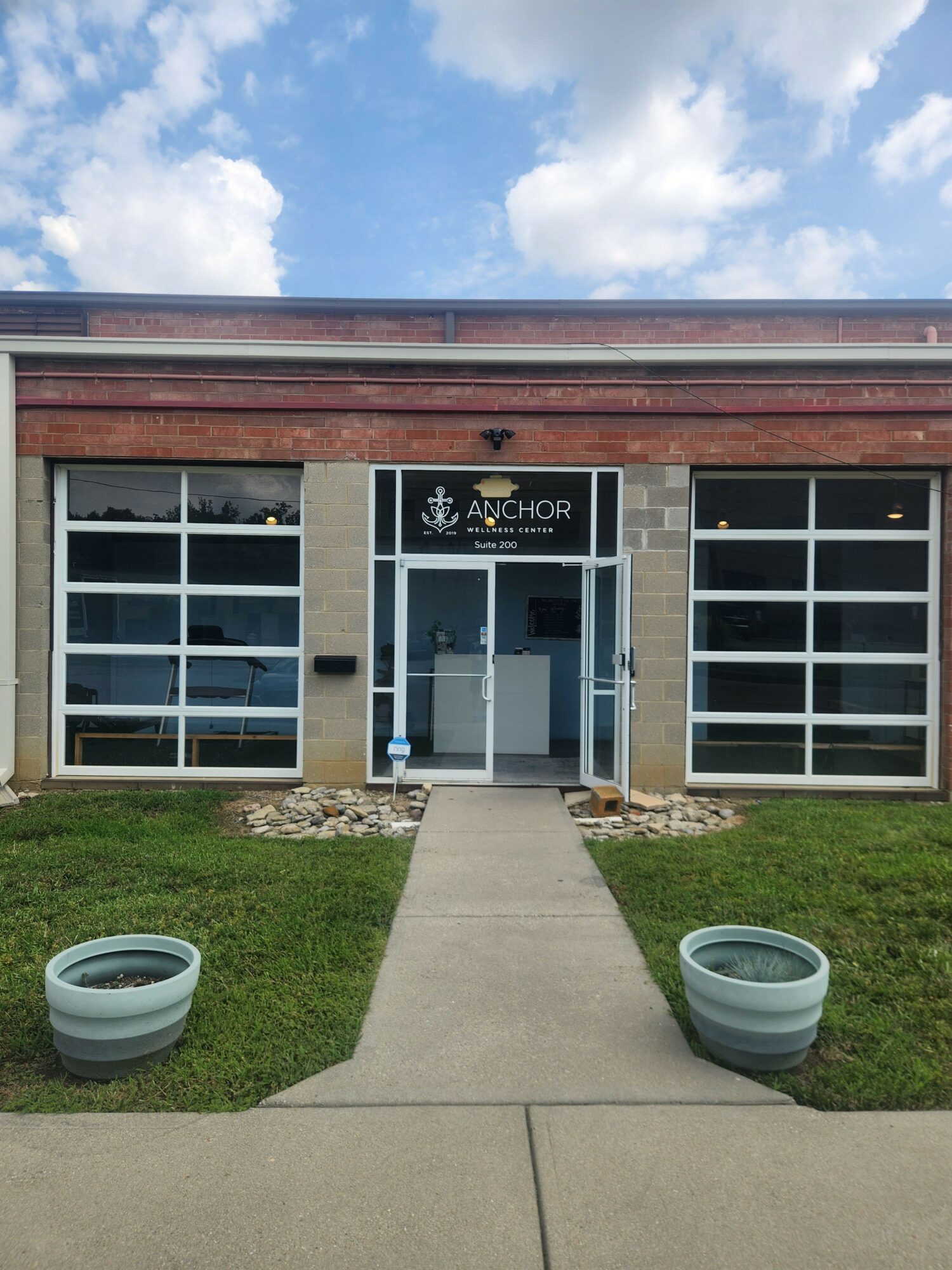Building entrance with glass door, windows, and two potted plants on a sidewalk, under a partly cloudy sky.