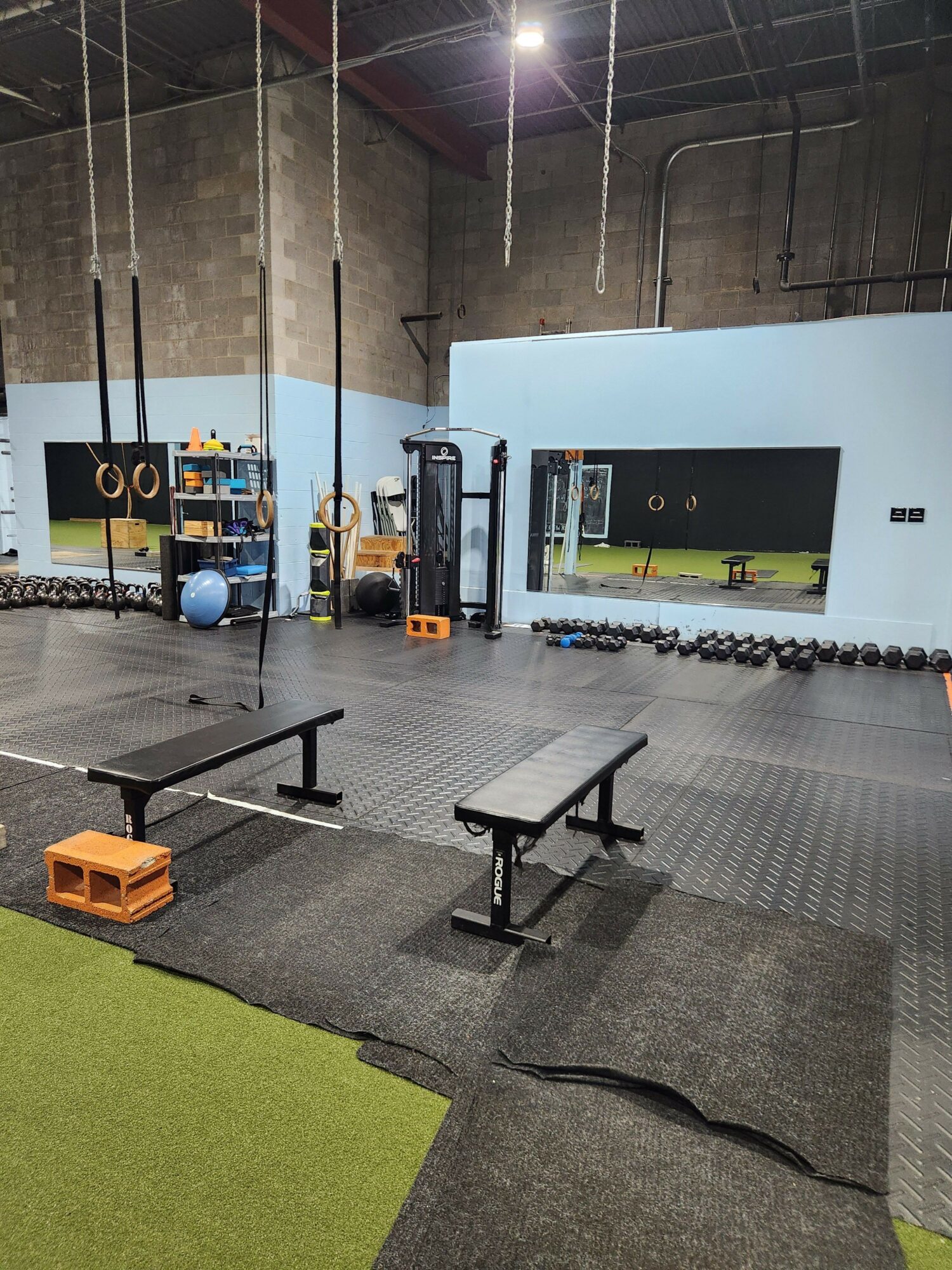 Indoor gym with workout equipment, mats, and a mirror, viewed from the side, with a green mat in the foreground.