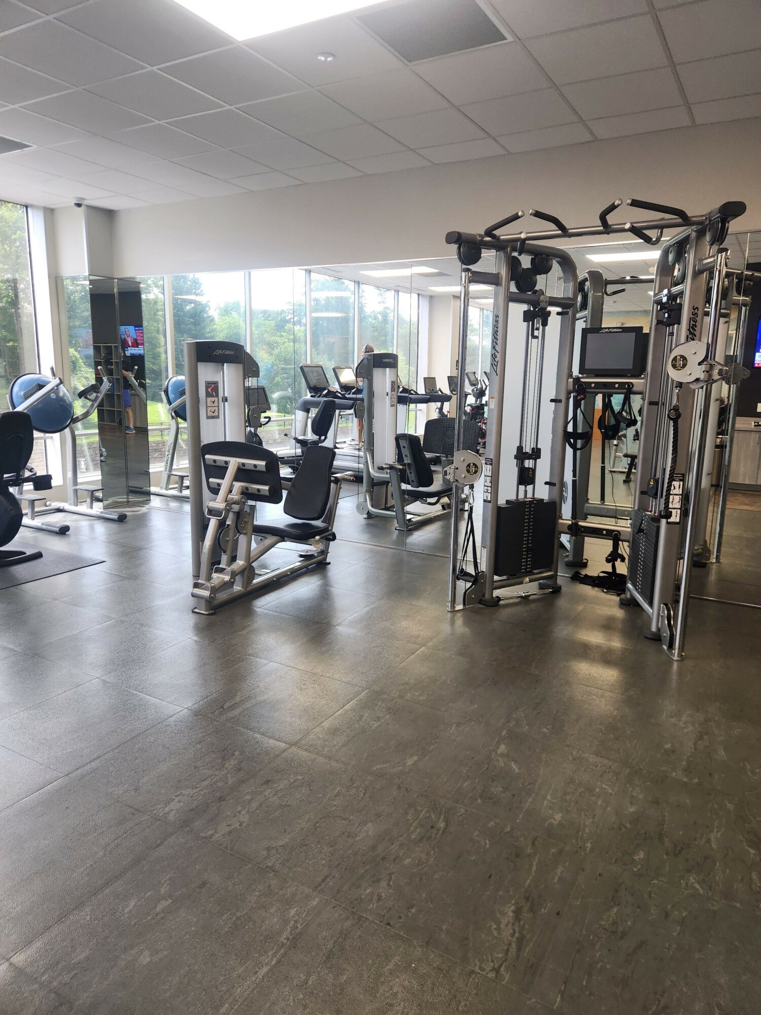 Gym with exercise equipment, large windows, and a polished floor, viewed from the side. Bright and spacious interior.