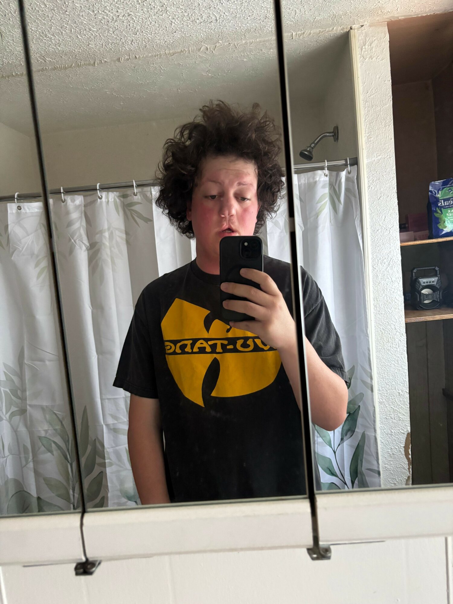Person with curly hair taking a mirror selfie in a room, wearing a black T-shirt with yellow logo, holding a phone.