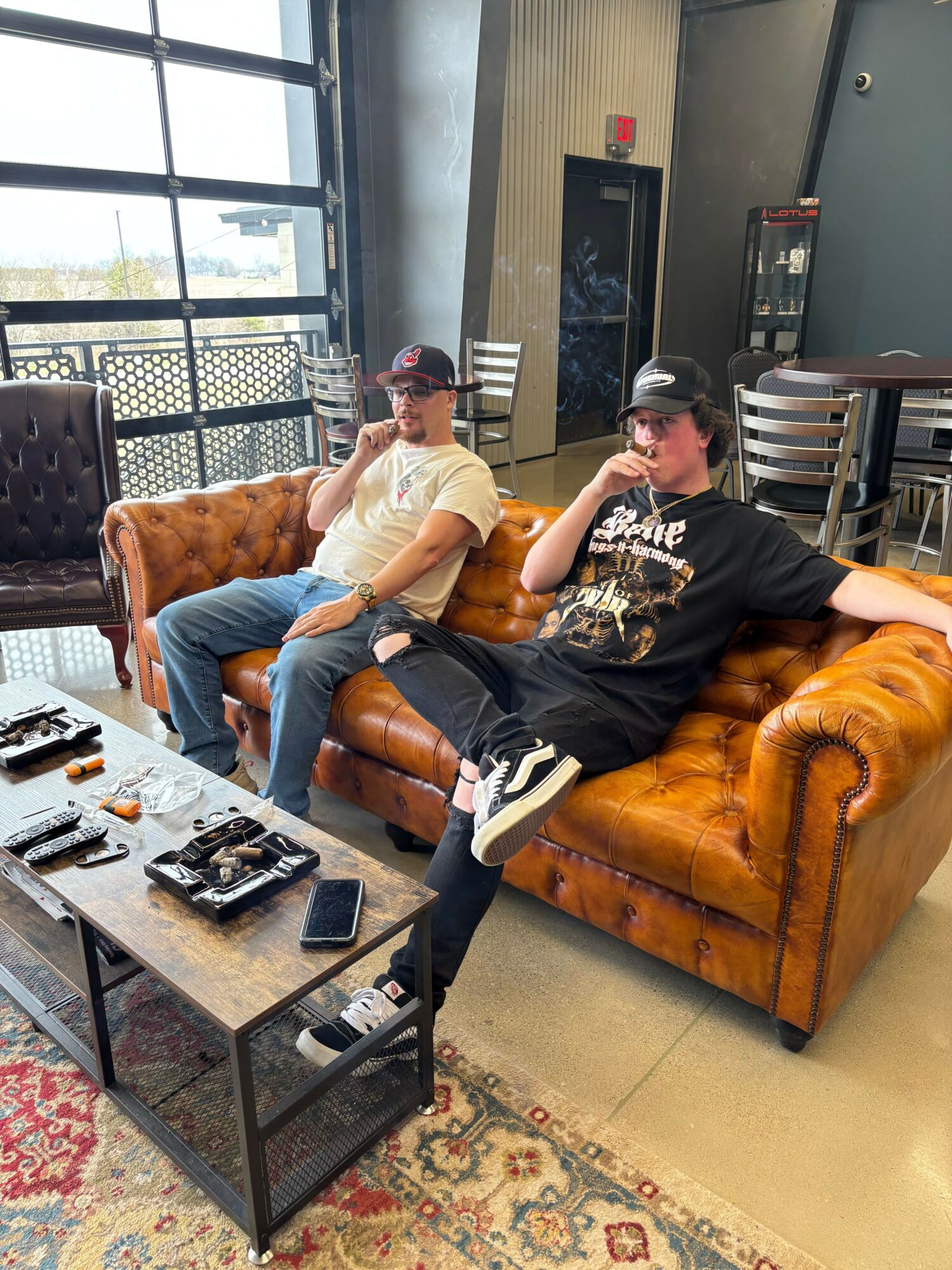 Two people sit on a leather couch in a modern room, one using a phone, with a coffee table and large window.