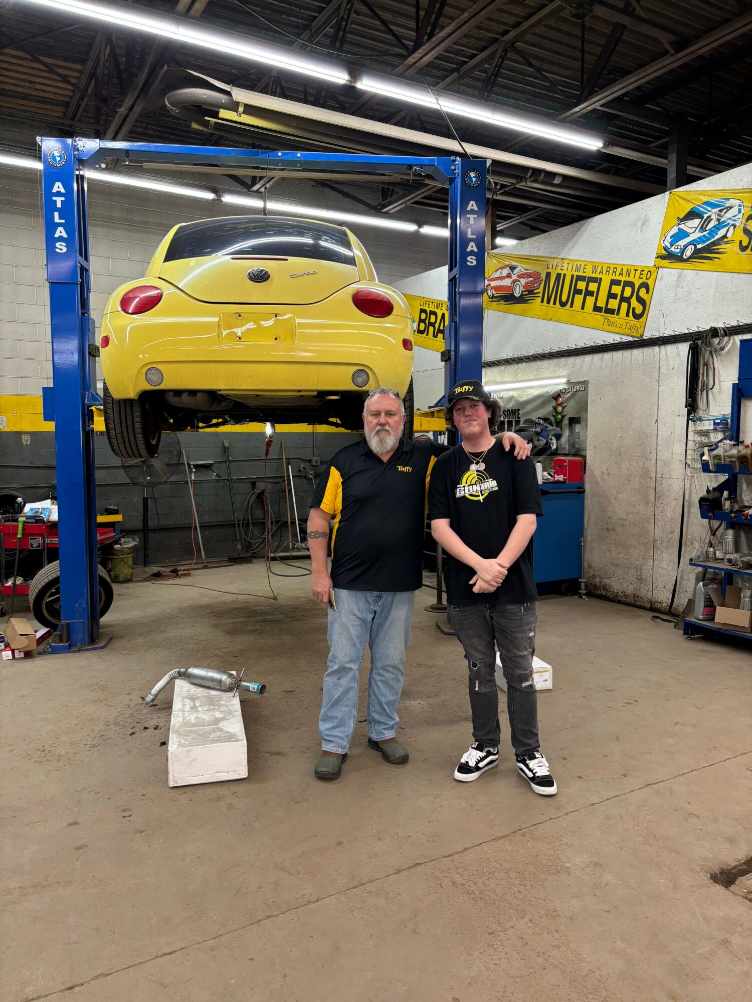 Two people standing next to a yellow car at a service station, with one person holding a yellow object, under a blue car lift.