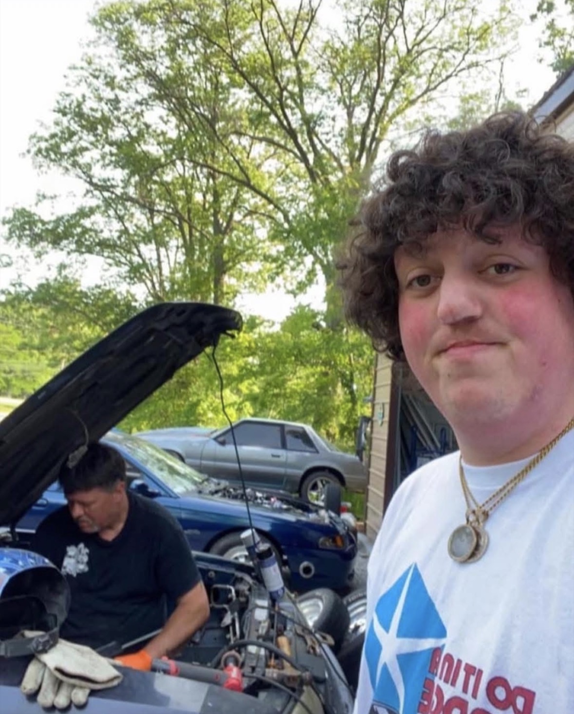 Person with curly hair smiling outdoors, car with open hood in background, trees, and another person working on a vehicle.