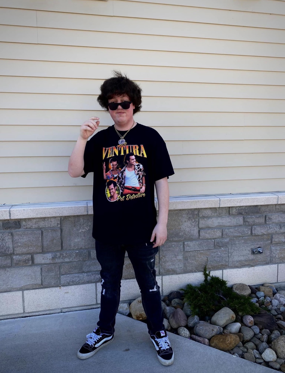 Person with curly hair wearing sunglasses, a black graphic T-shirt, and jeans, standing outdoors in front of a beige wall.