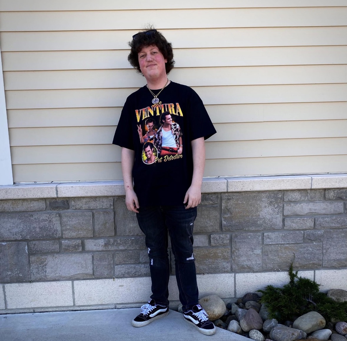 Person with curly hair wearing a black graphic T-shirt, dark jeans, and sneakers, standing against a beige wall with stone base.