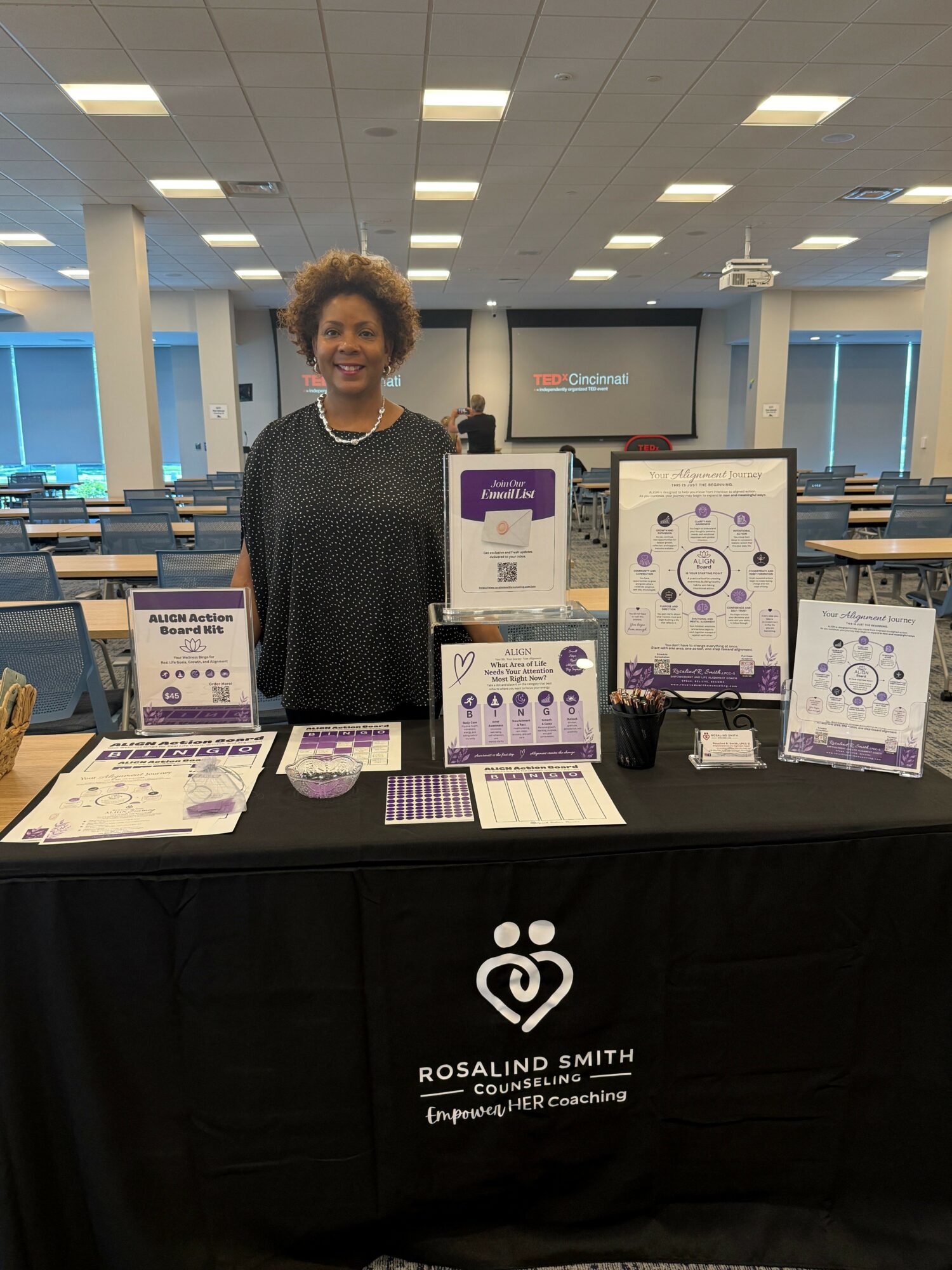 Person standing behind a table with informational posters and materials, in a well-lit indoor space.