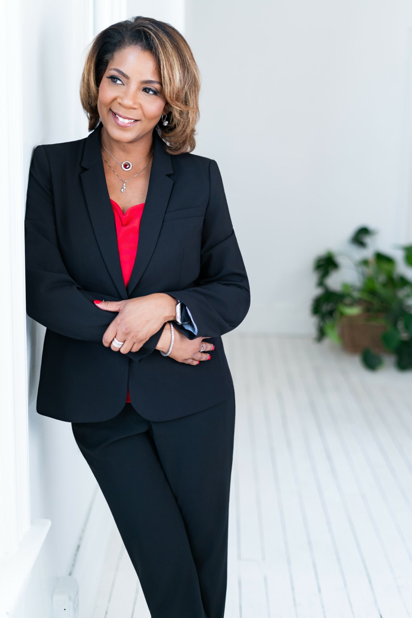 Woman in black suit with red top, smiling, standing indoors near a white wall and potted plant.