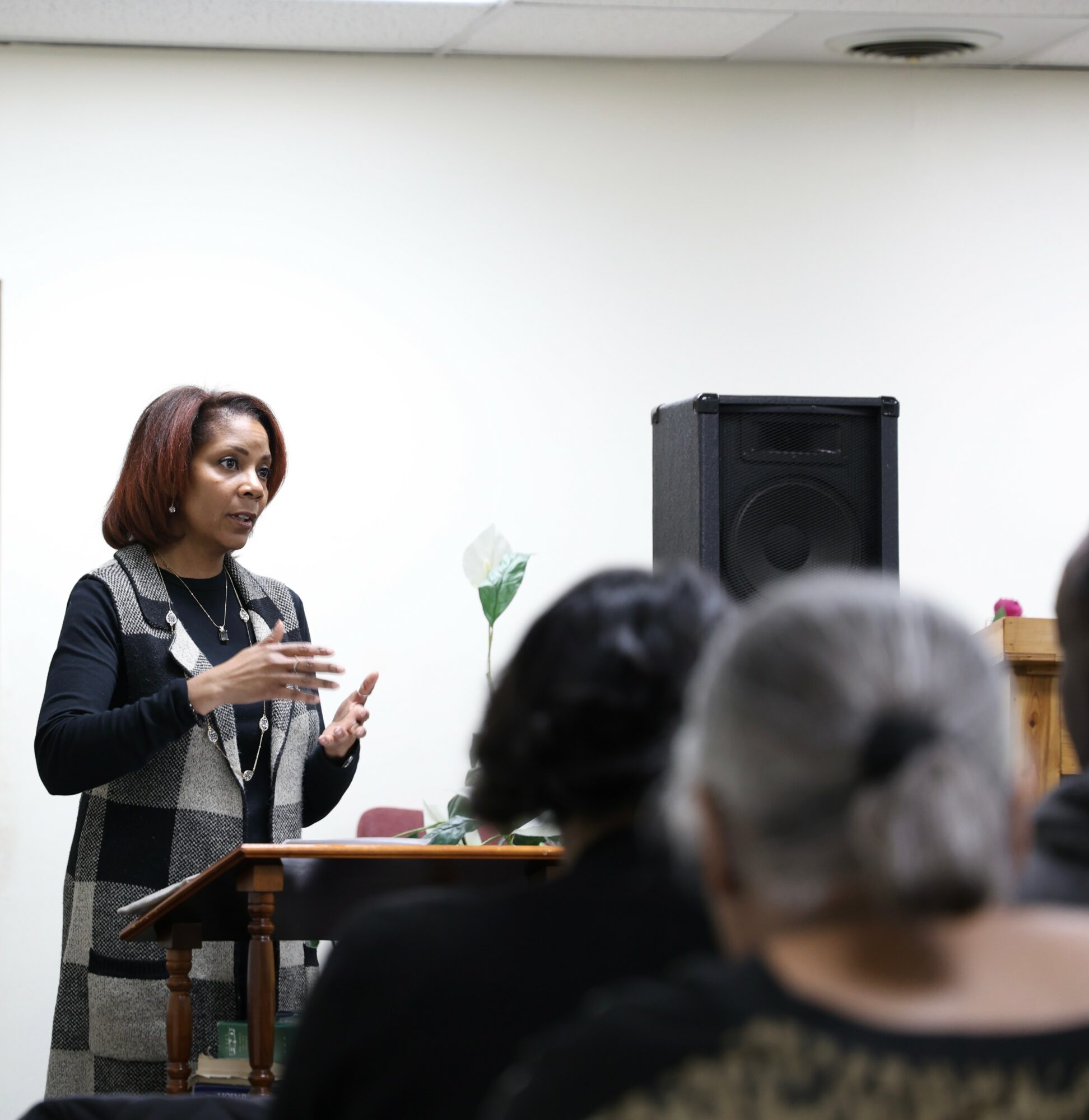 Woman speaking to an audience in a room with a speaker and a table, audience members visible from behind.