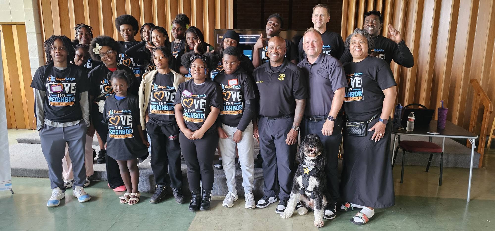 Group of people standing indoors, some wearing matching shirts, with a dog sitting in front, wooden wall background.