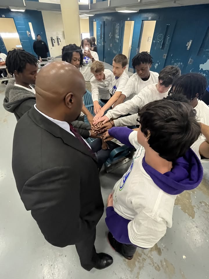 Group of students and a man with a bald head and suit in a school hallway, hands stacked together, diverse group, lockers in background.
