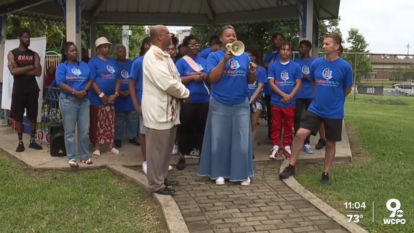 Group of people wearing blue shirts gathered outdoors, some listening and others speaking, with a man holding a microphone.