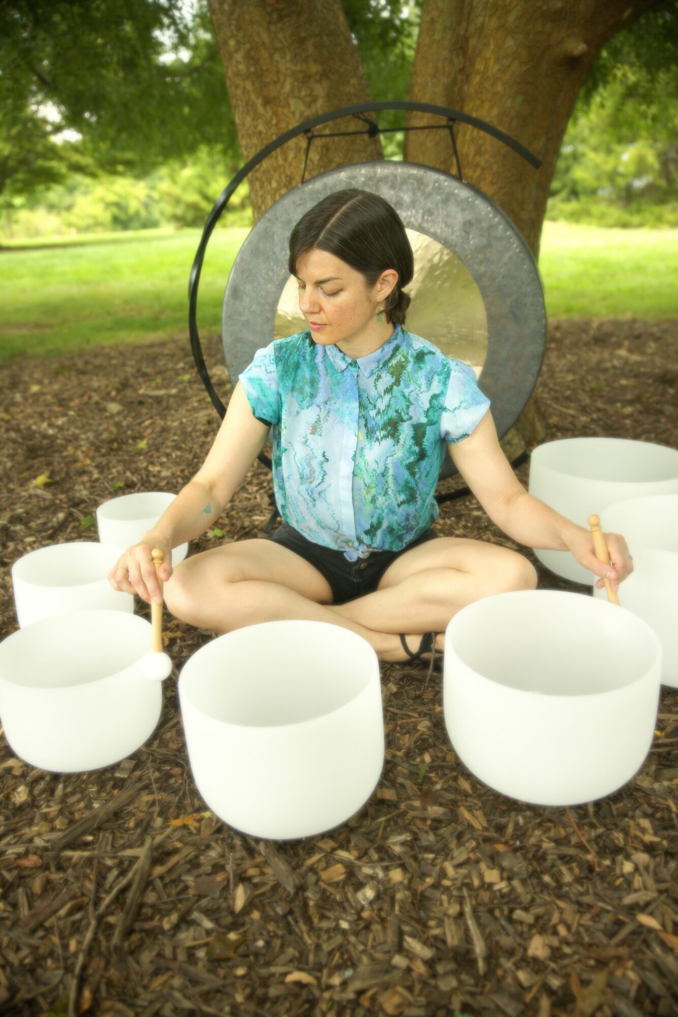 Young woman sitting cross-legged outdoors with white bowls around her, a large tree in the background, and a circular gong behind her.