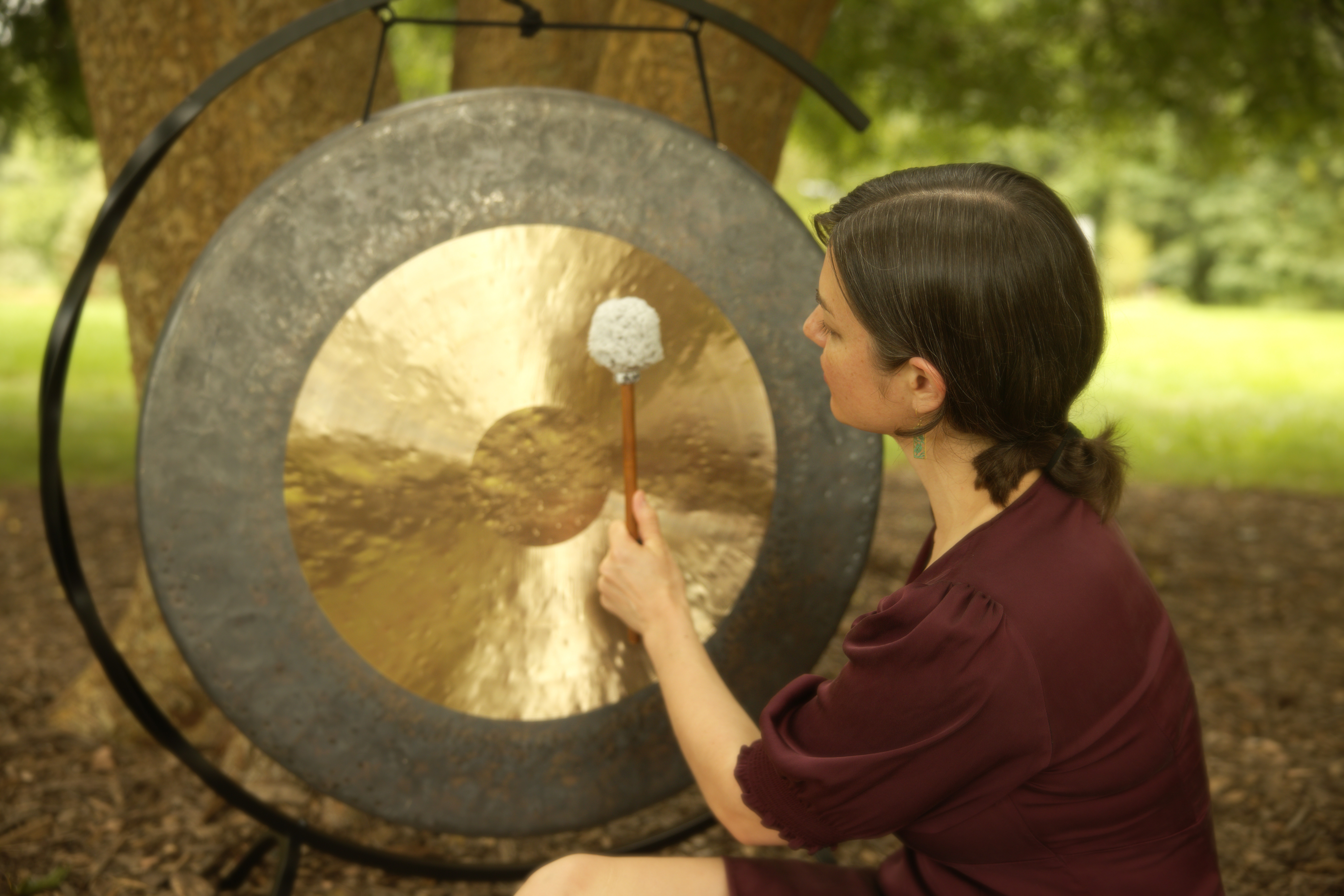 Woman strikes a gong with a mallet outdoors, surrounded by trees and greenery.
