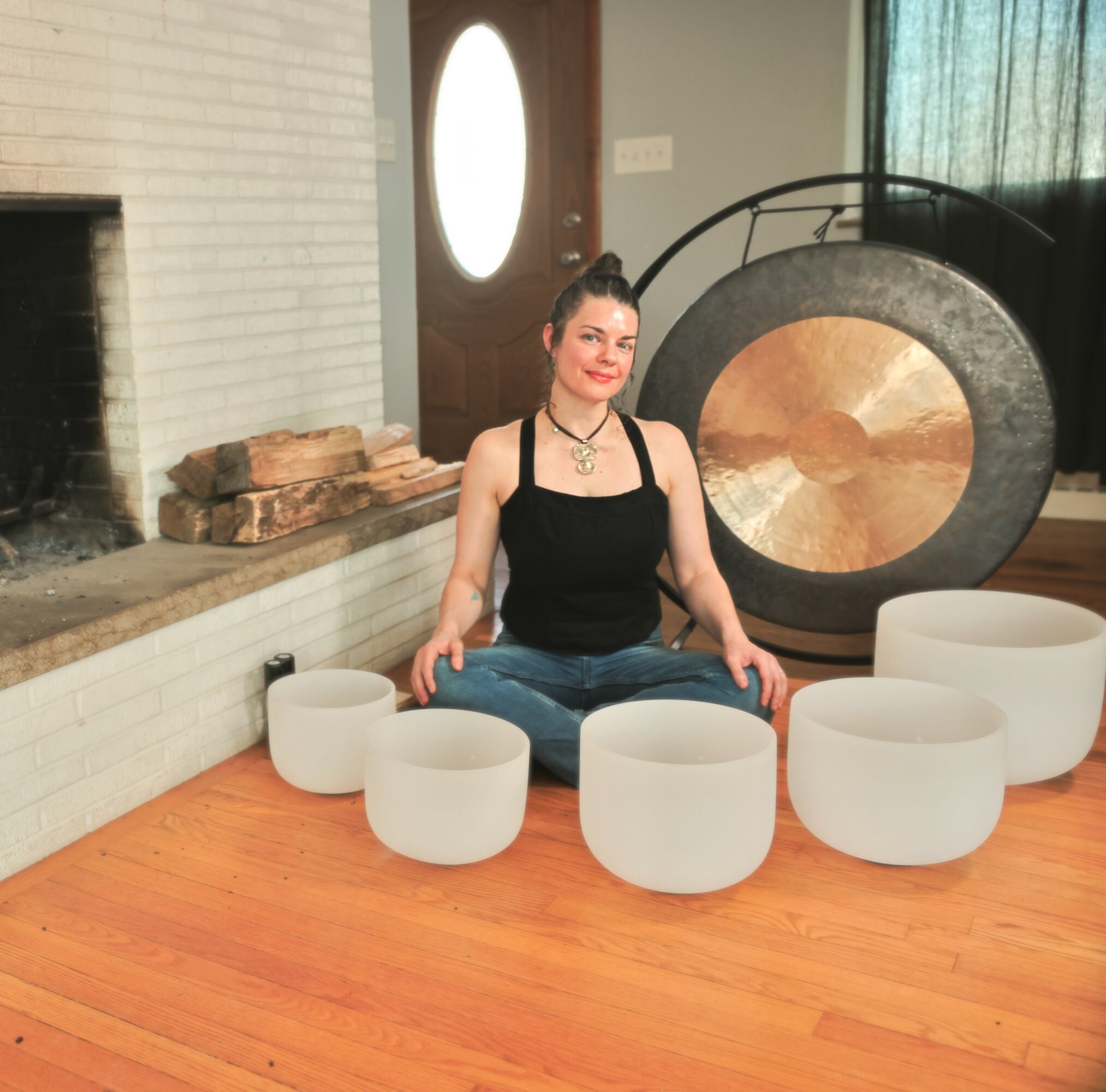 Person sitting cross-legged on wooden floor surrounded by white crystal singing bowls, with a large gong and fireplace in background.