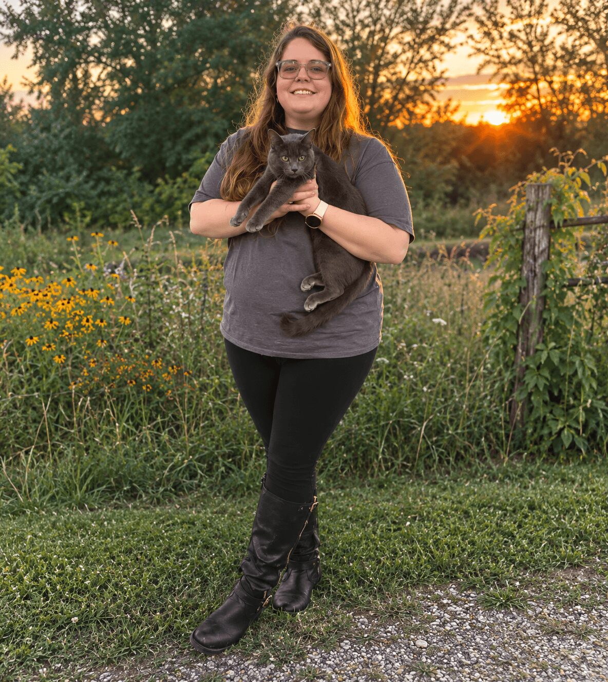 Woman holding a cat outdoors during sunset, standing on grass with trees and a fence in the background.