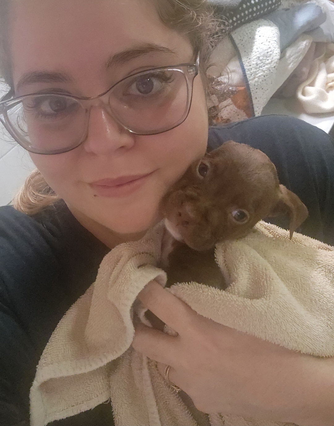 Person with glasses holding a small brown puppy wrapped in a towel, indoors with clothes in the background.