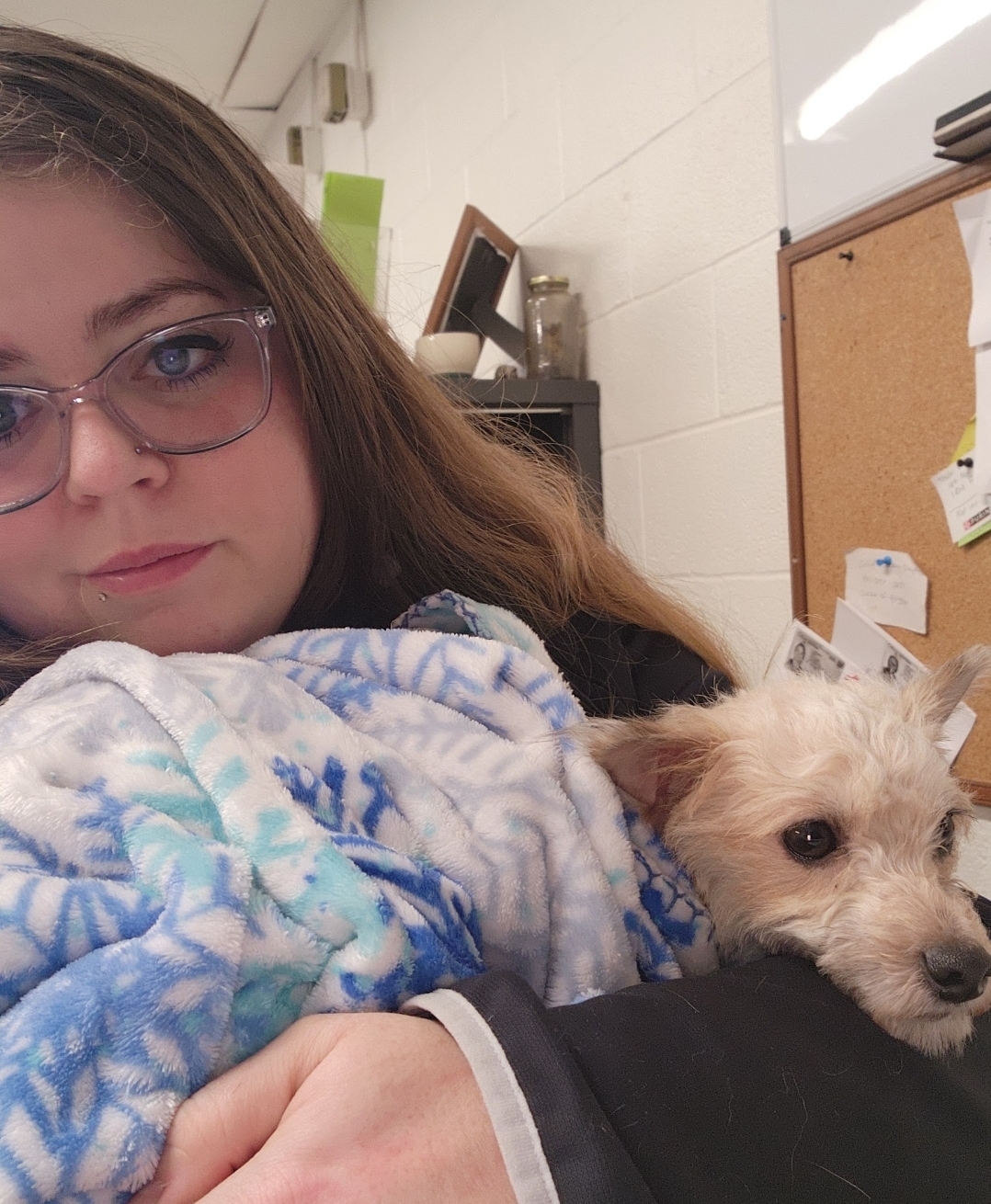 Young woman with glasses holding a small dog wrapped in a blue and white blanket in an indoor setting.