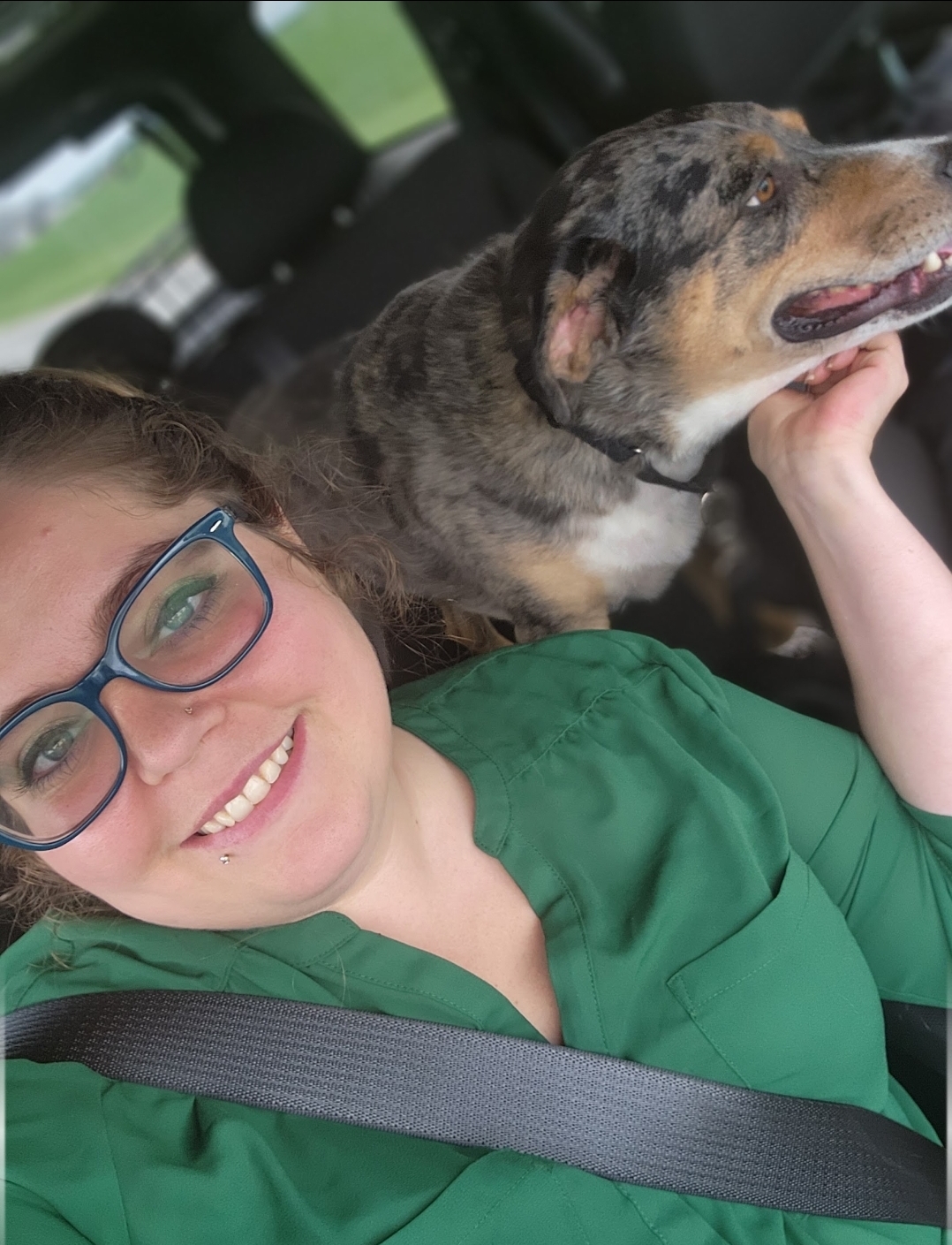 Person with glasses smiling in a car, holding a dog with a brindle coat and tan markings, both looking happy.