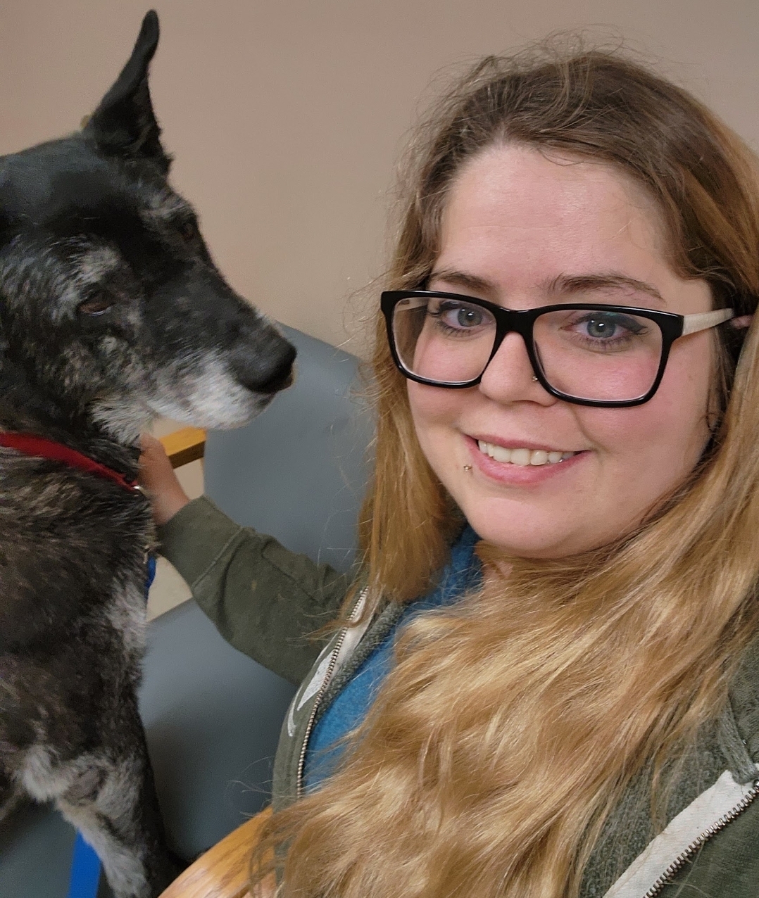 Young woman with glasses smiling next to a black and white dog, both facing each other, indoors.