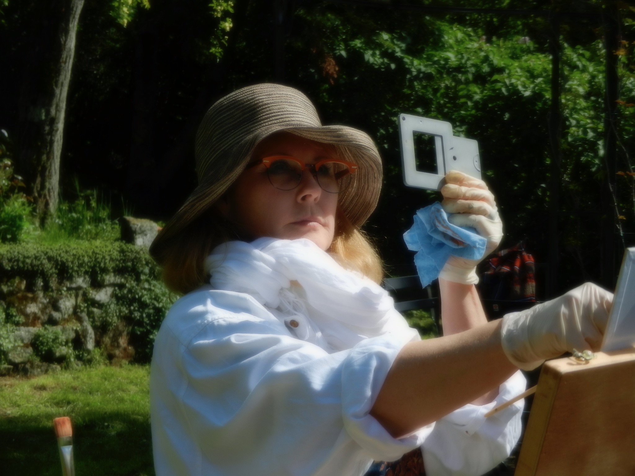 Woman wearing a hat and sunglasses outdoors, holding a camera, with trees in the background.