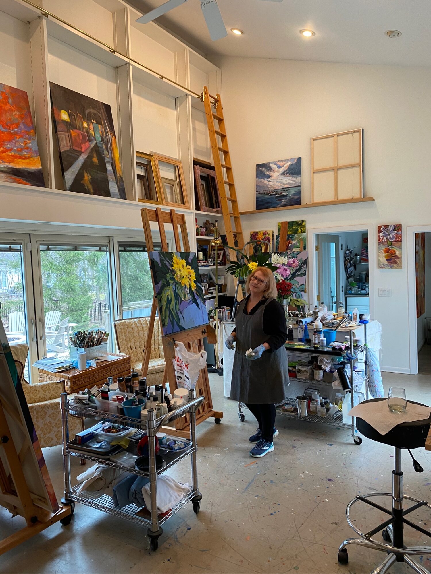 Two women in an art studio surrounded by paintings, art supplies, and large windows, engaging in conversation.