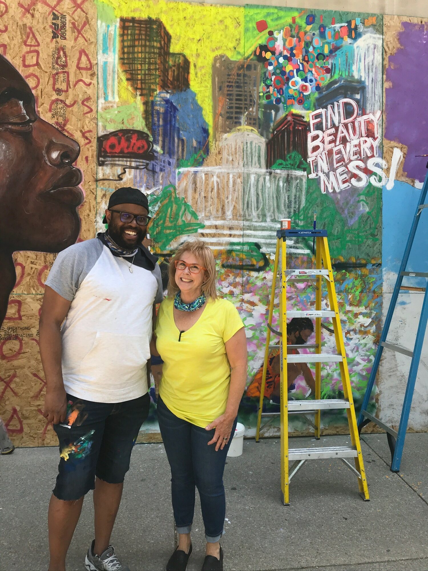 Two people stand in front of a colorful mural with a ladder nearby, smiling at the camera.