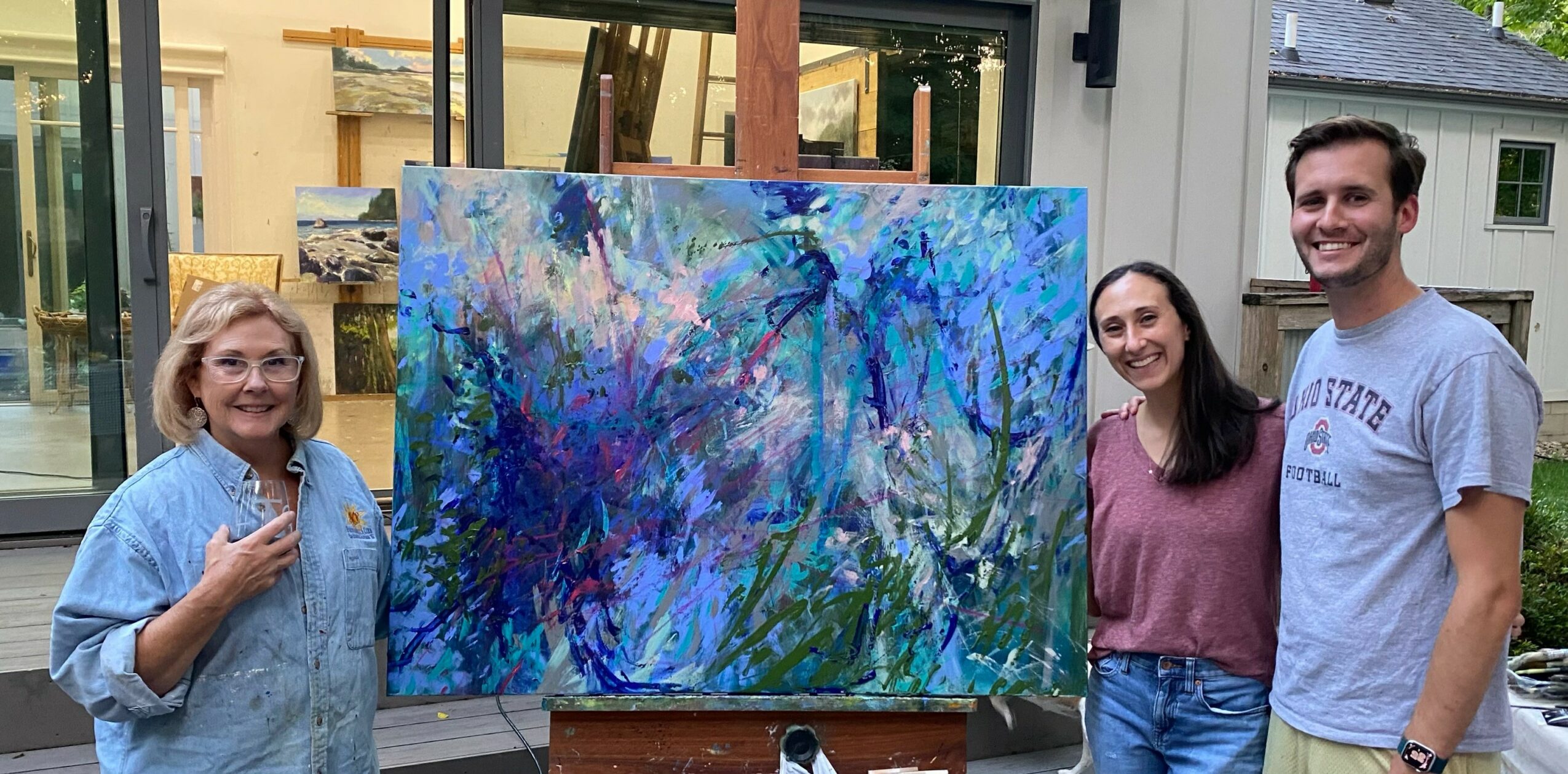 Three people standing outdoors next to an abstract painting on an easel, smiling, with buildings in the background.