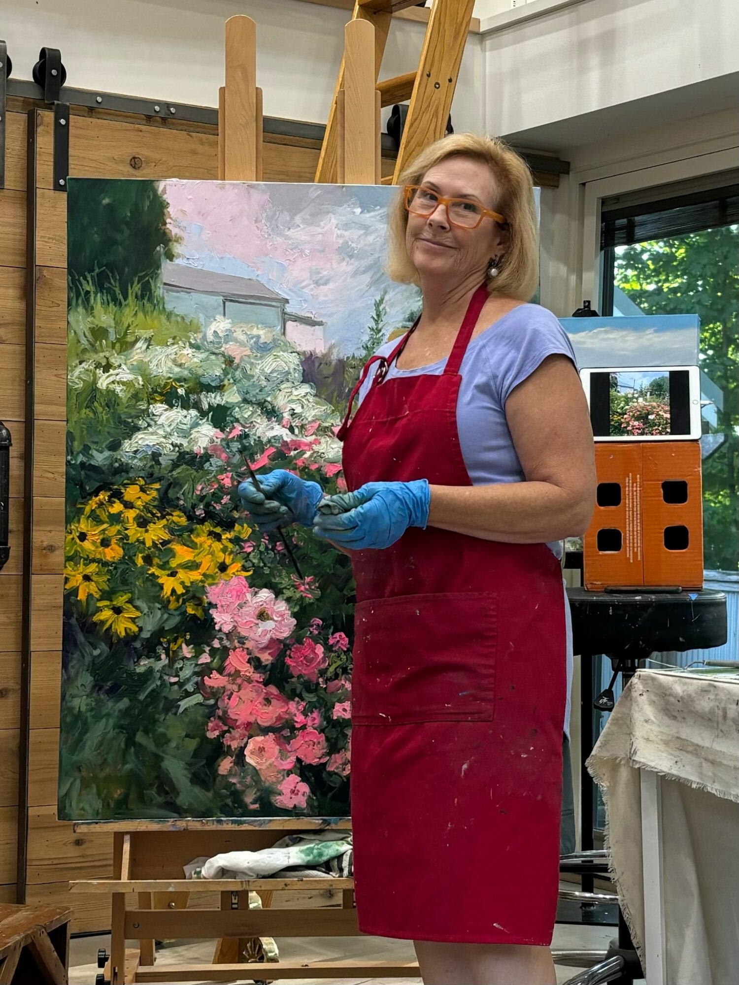 Woman in red apron and gloves painting a garden scene on canvas, smiling, in art studio.