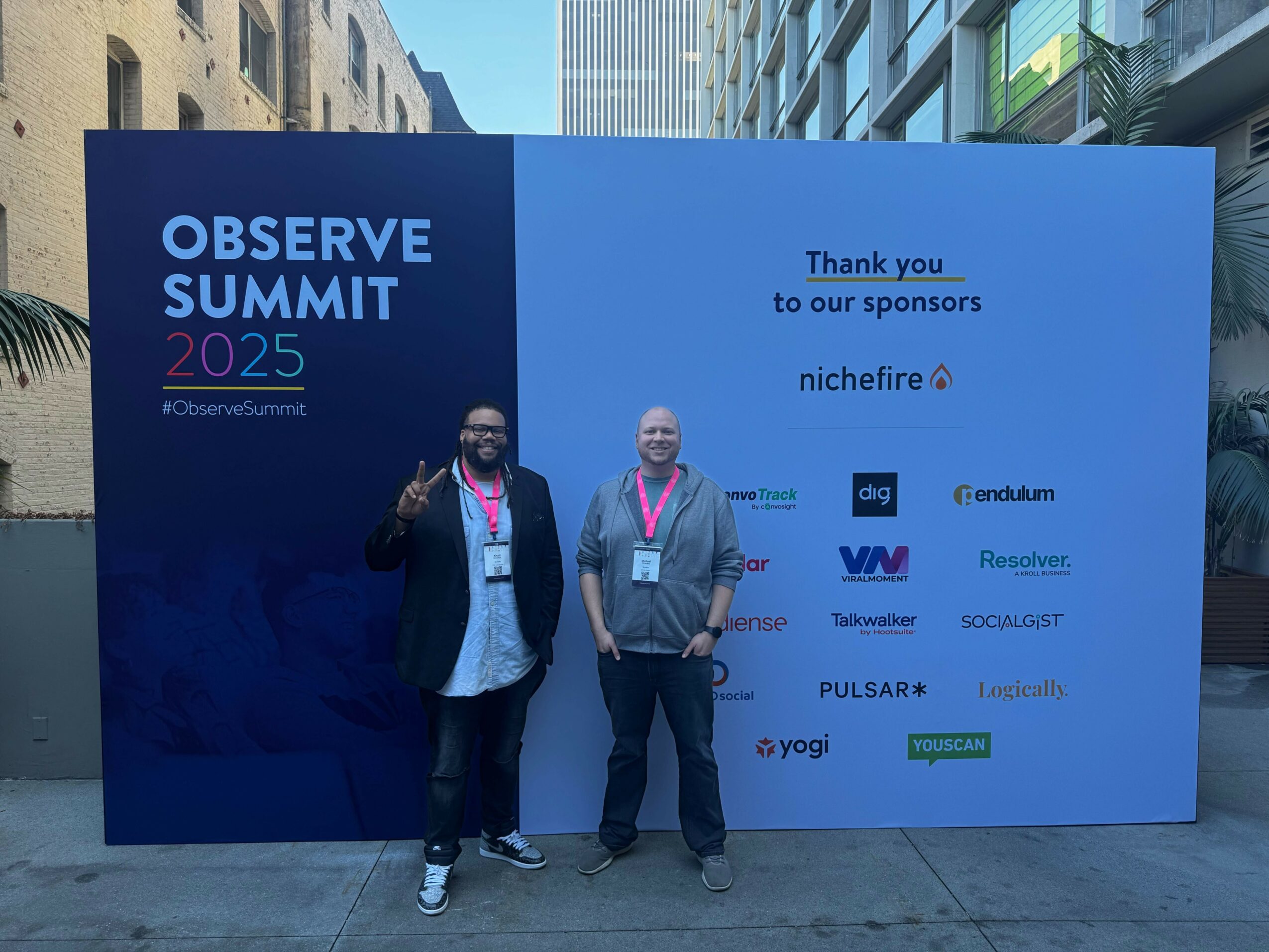Two men standing in front of a large blue and white event backdrop at Observe Summit 2025, outdoors in an urban area.