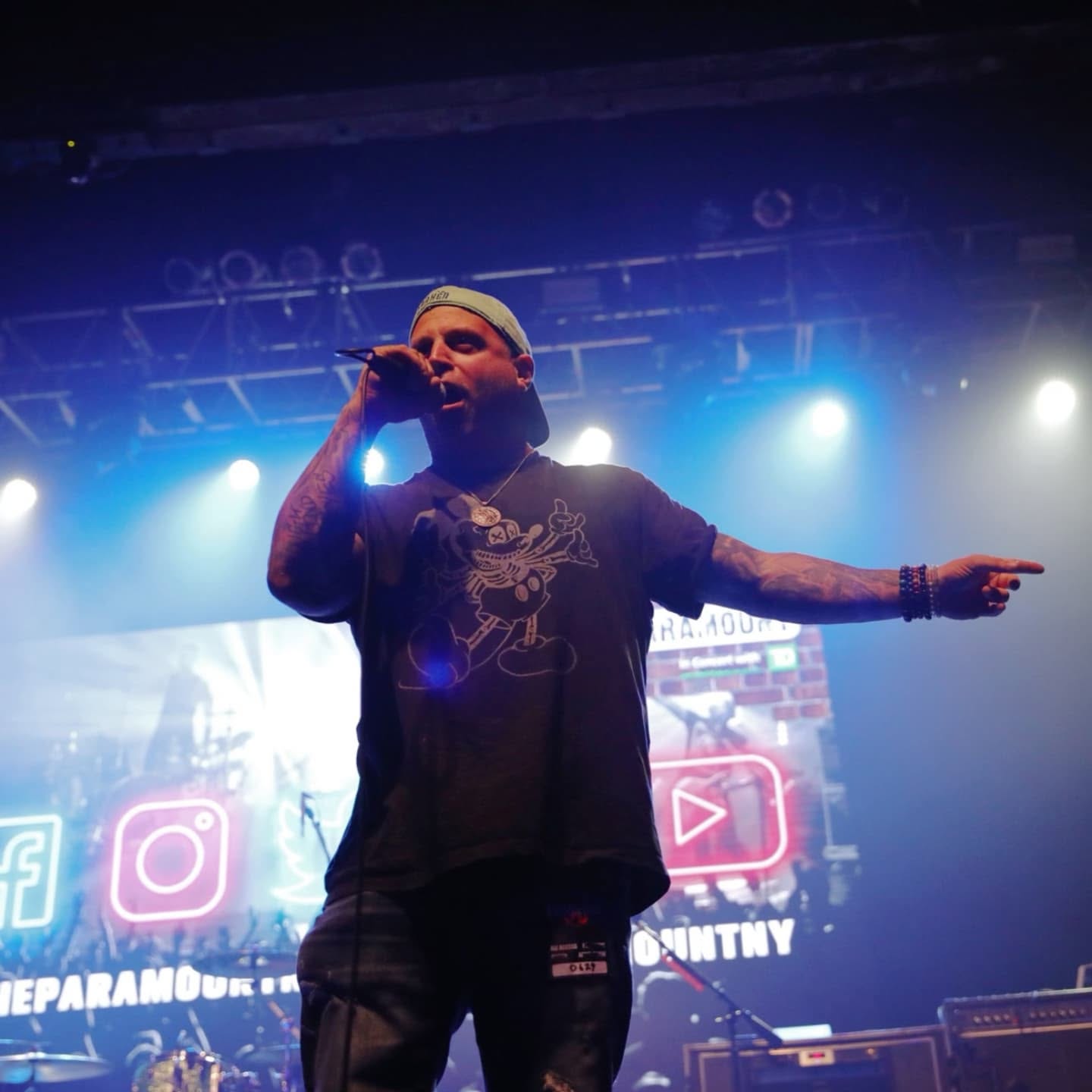 Performer singing into microphone on stage with bright lights and social media icons in background.