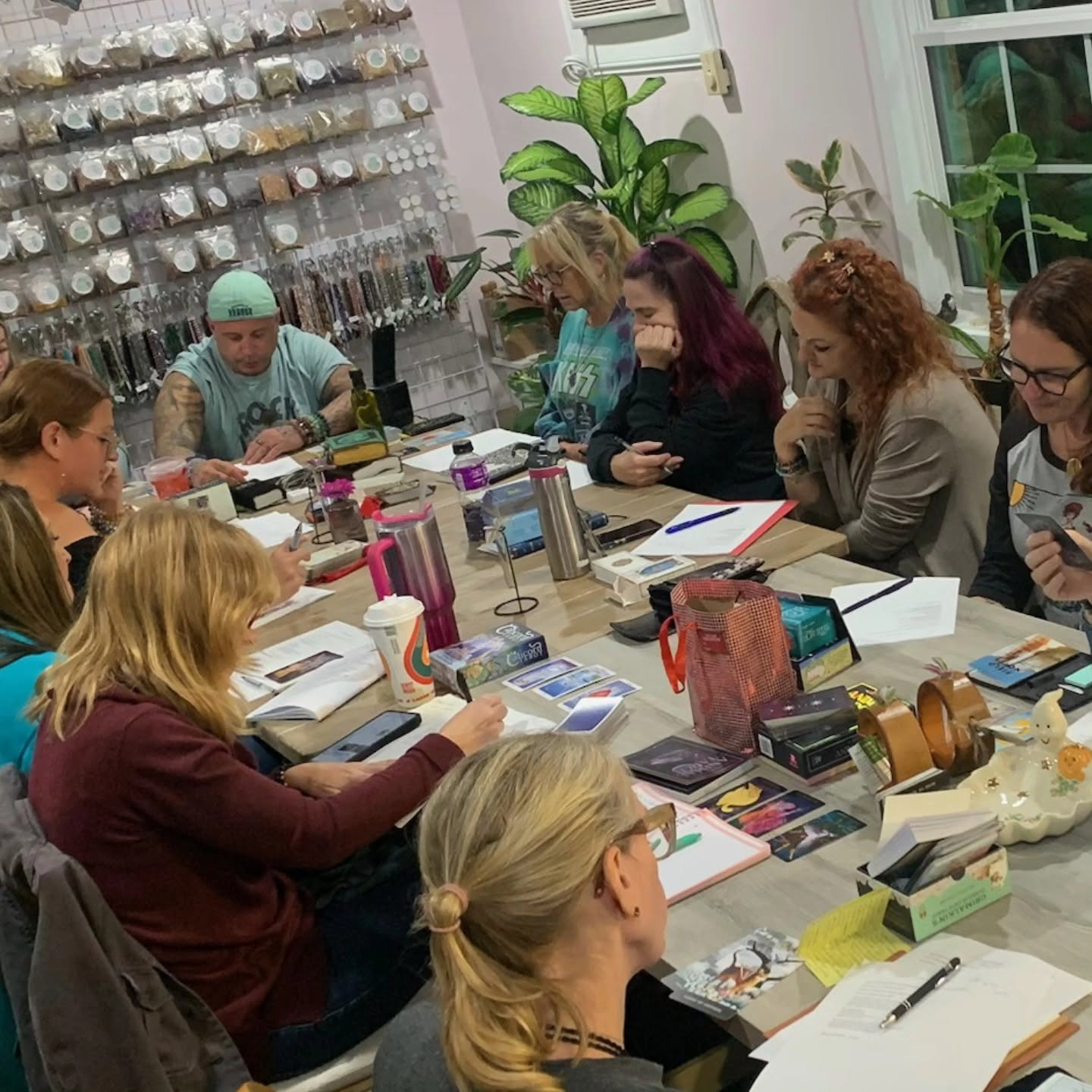 Group of women sitting around a table in a room with shelves of jars and plants, engaged in a discussion or workshop.