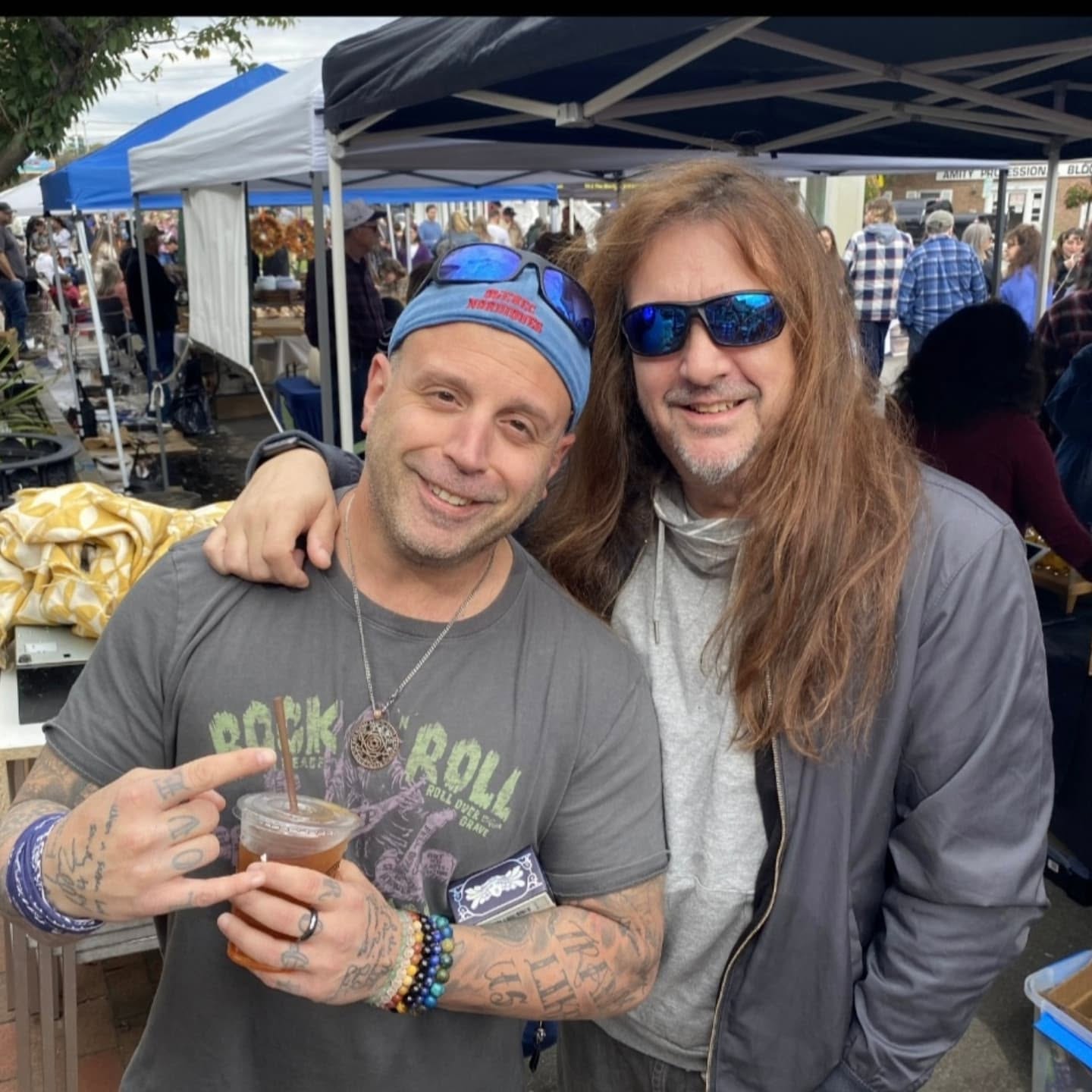 Two men smiling at an outdoor event, one with long hair and sunglasses, the other with a cap and tattoos, holding a drink.