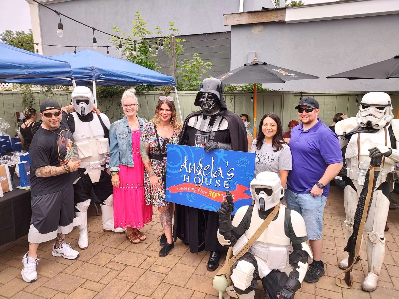 Group of people, including individuals dressed as Star Wars characters, standing outdoors with tents and umbrellas, smiling and posing for a photo.