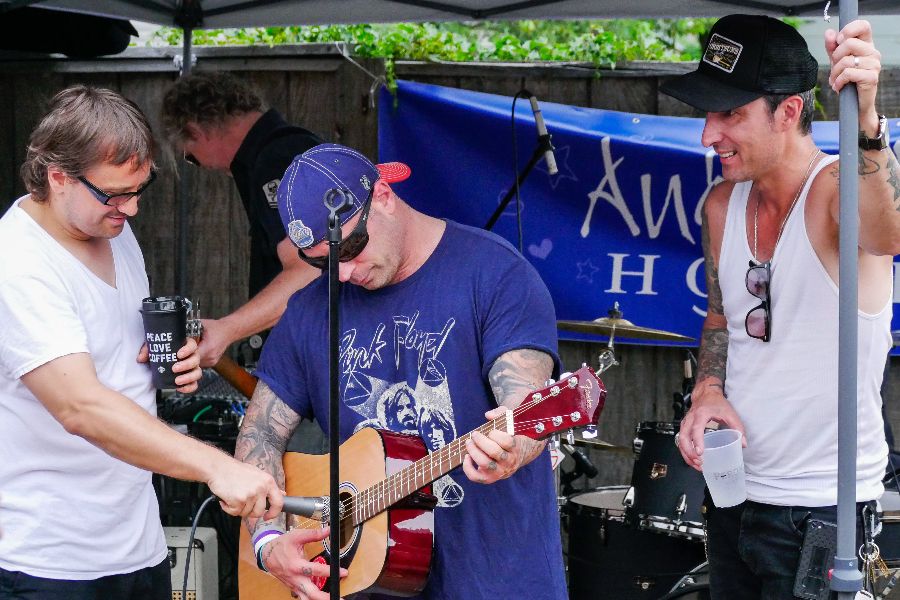 Three men playing guitar and singing outdoors under a canopy, with a blue banner in the background.